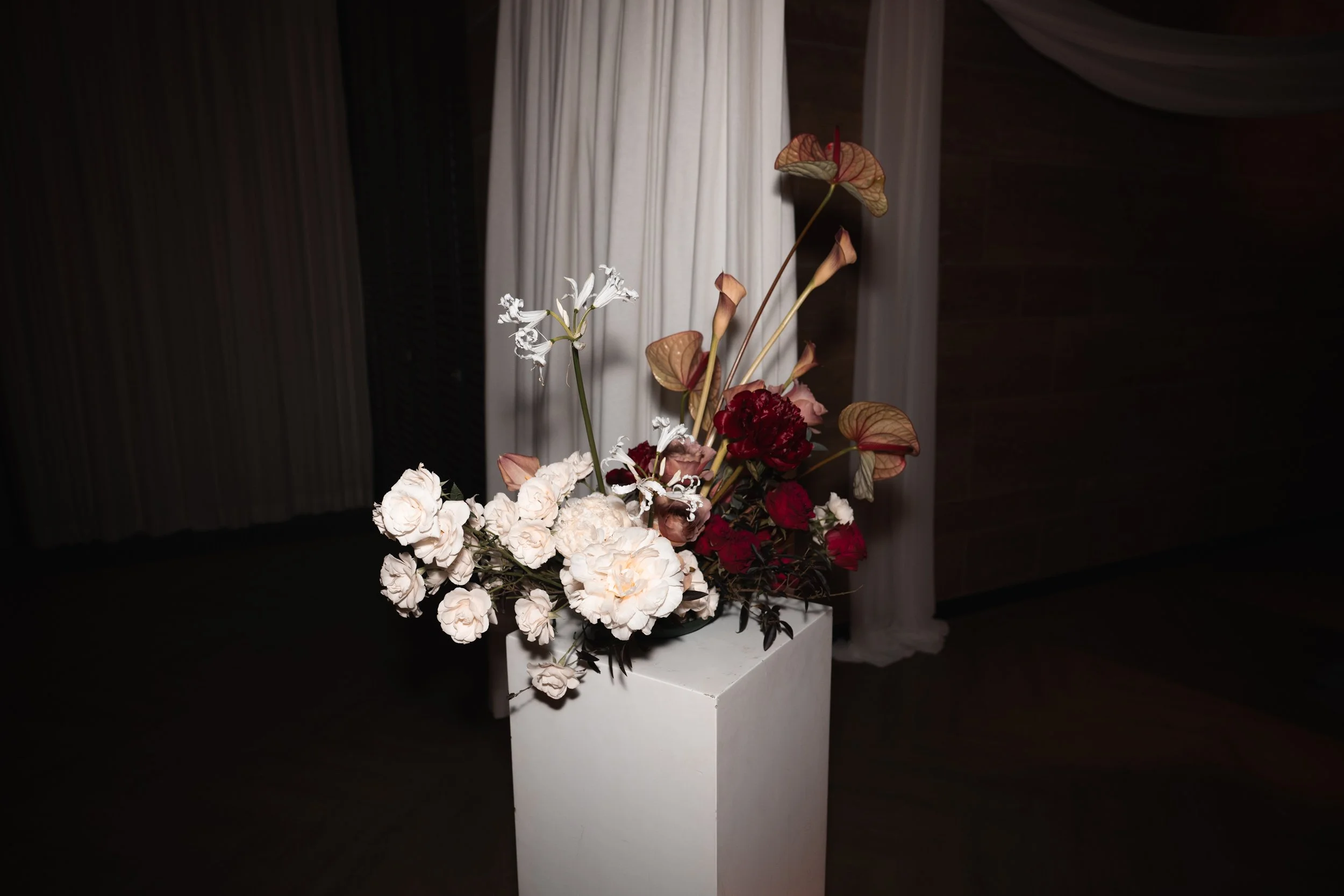 A floral arrangement with white, red, and pink flowers in a white vase, set against a dark background with white curtains.