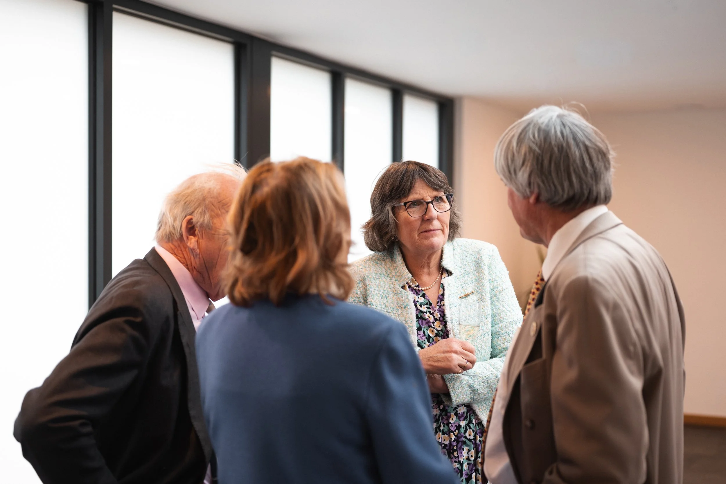 A group of four older adults engaged in a conversation indoors near large windows.