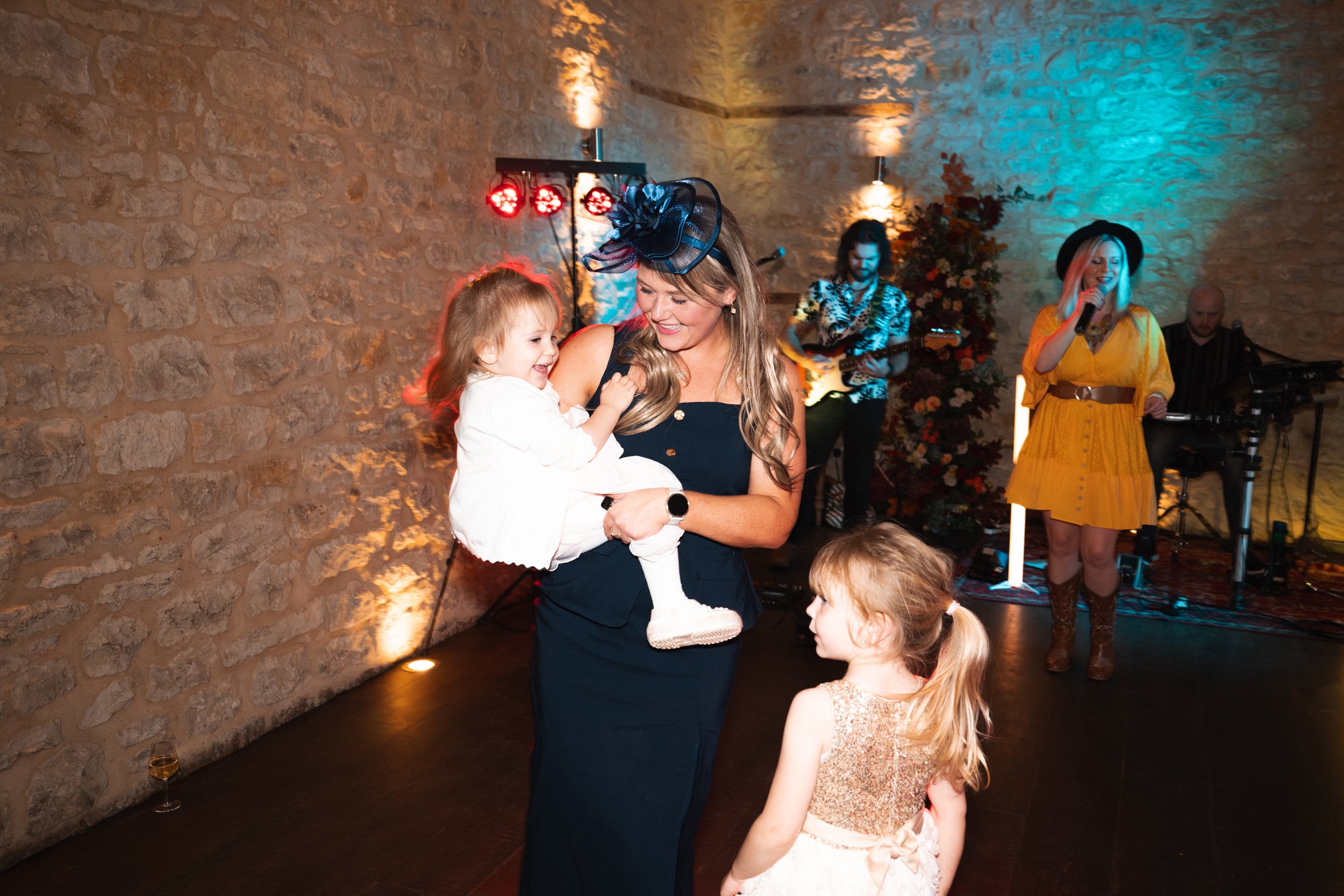 Woman in dark dress holding a laughing young girl, with another girl standing nearby, at a lively indoor party with a live band playing on the stage.
