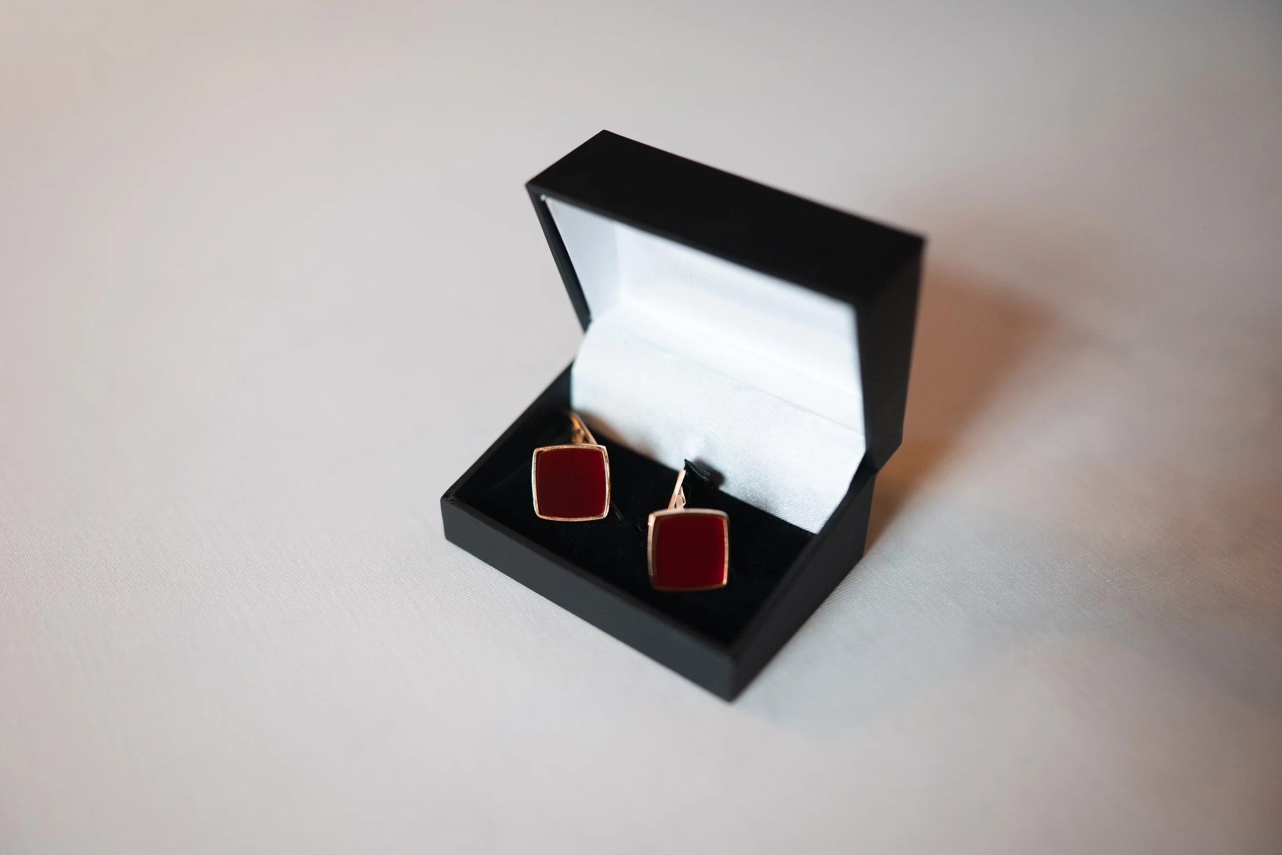 A pair of red square cufflinks in an open black jewelry box with a white interior, placed on a white surface.