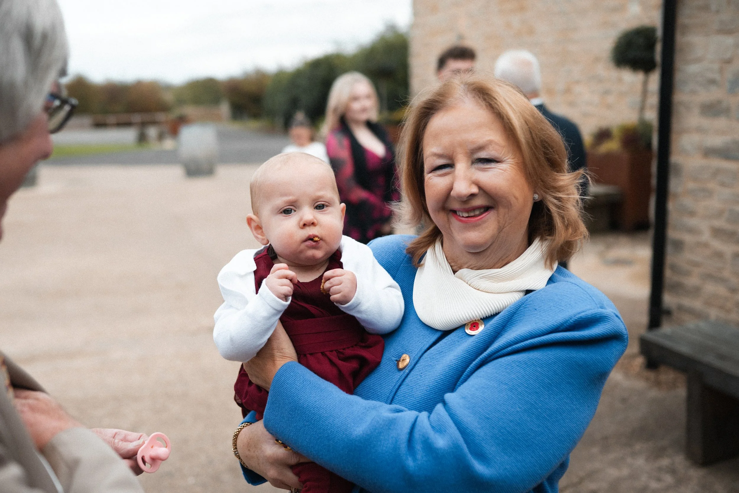 A woman with red hair and a blue jacket holding a baby girl in a maroon dress outside. Other people are in the background near a stone building.