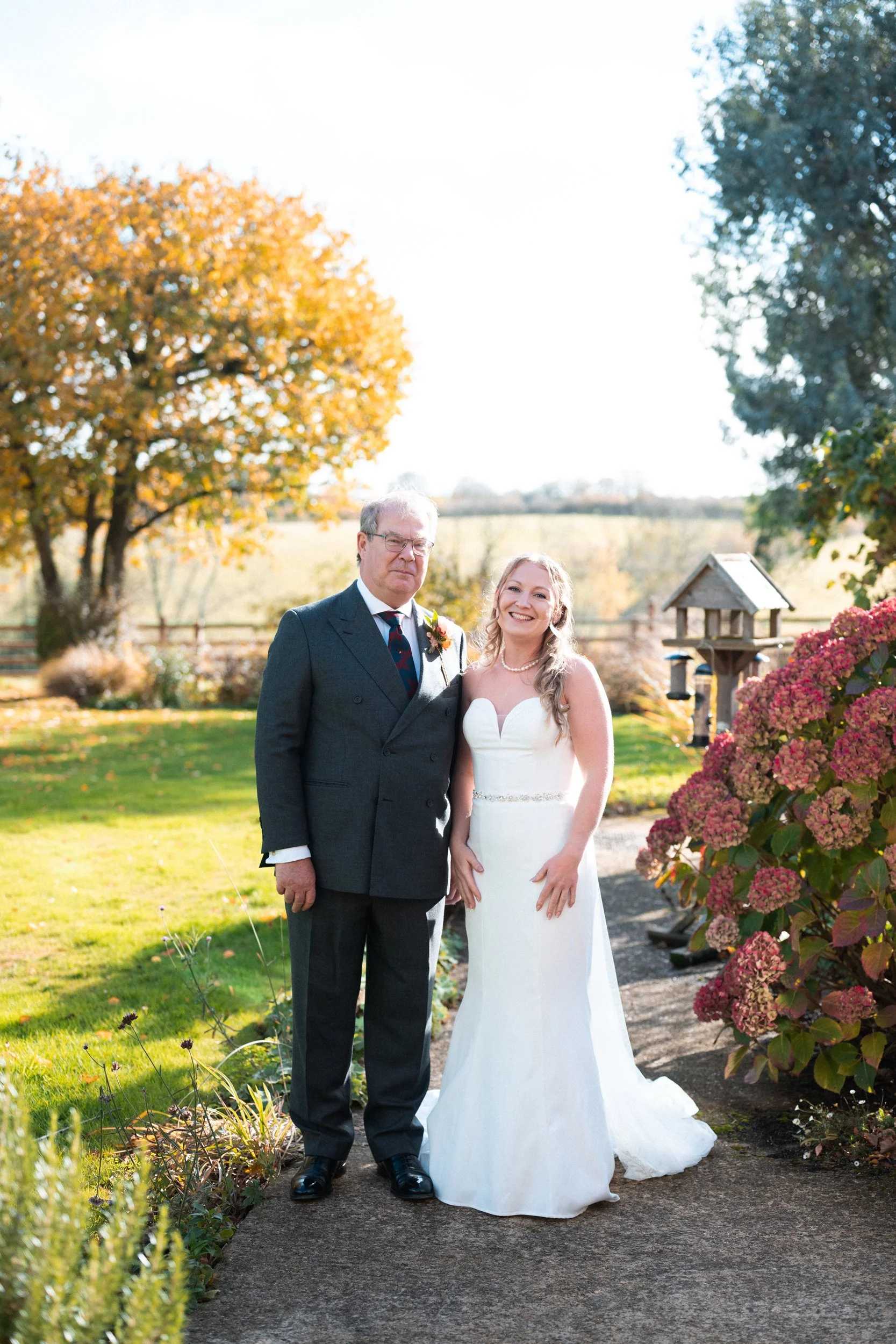 A bride in a white wedding gown standing next to a man in a dark suit outdoors on a sunny day, with trees and colorful autumn foliage in the background.
