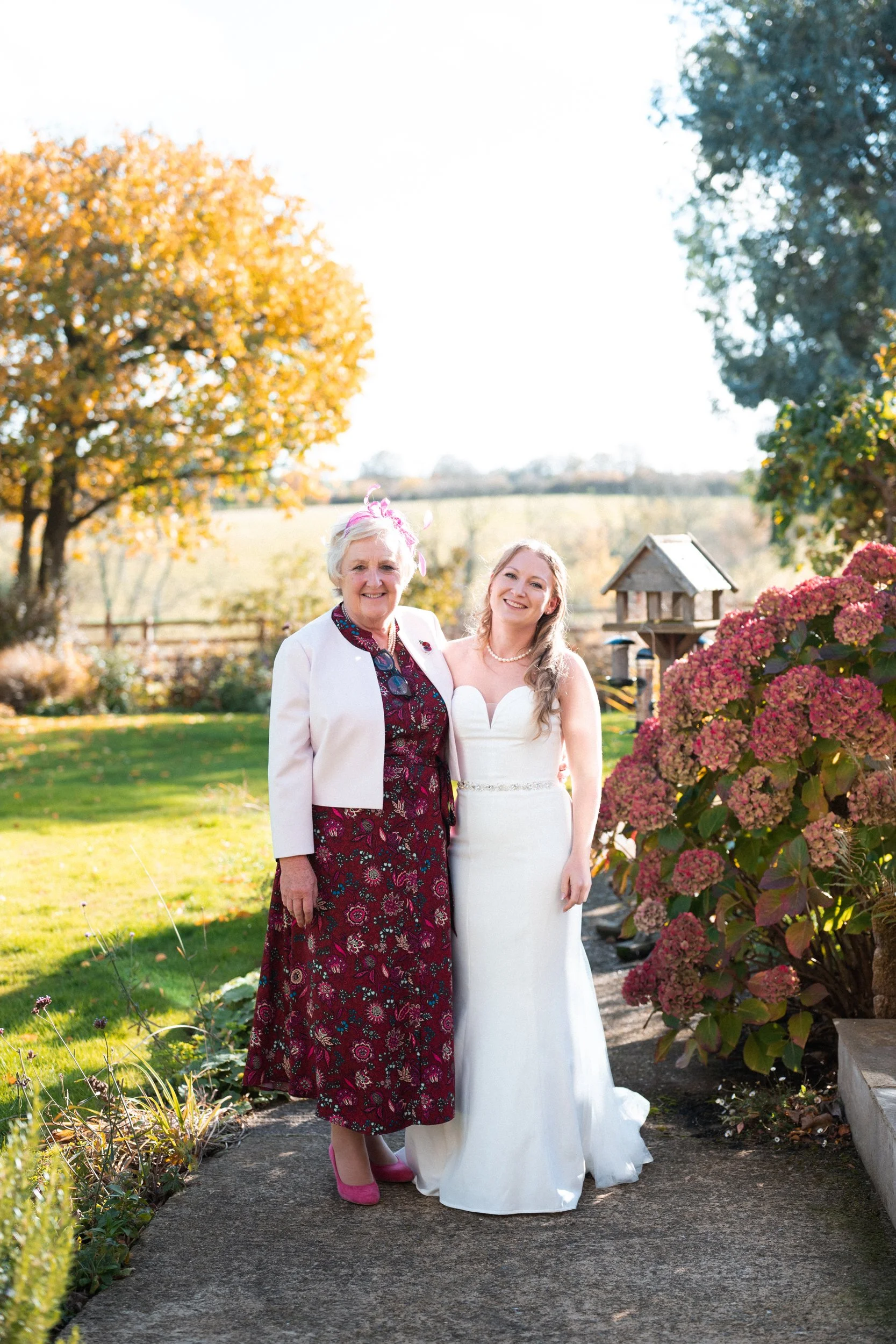 A bride and her mother standing outdoors on a sunny day, smiling. The bride is wearing a white wedding gown, and the mother is in a burgundy floral dress with a white jacket. There are trees and pink hydrangeas in the background.