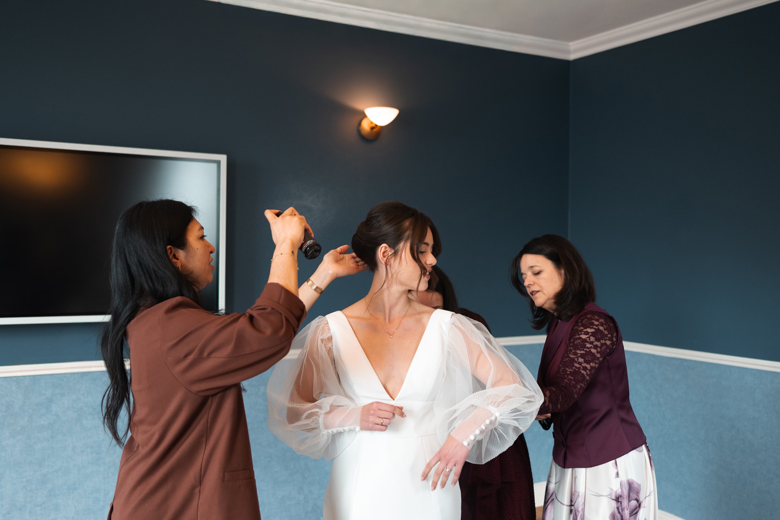 A bridal woman in a white gown with sheer puffed sleeves being assisted by women, one holding her hair and the other adjusting her dress, in a room with blue walls and a wall-mounted TV.
