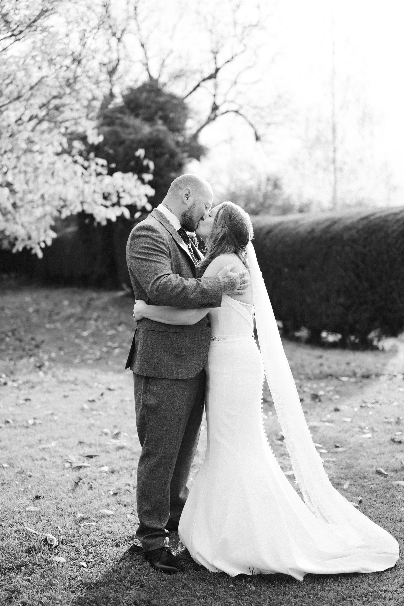 A black and white photo of a bride and groom sharing a kiss outdoors, with trees and bushes in the background.