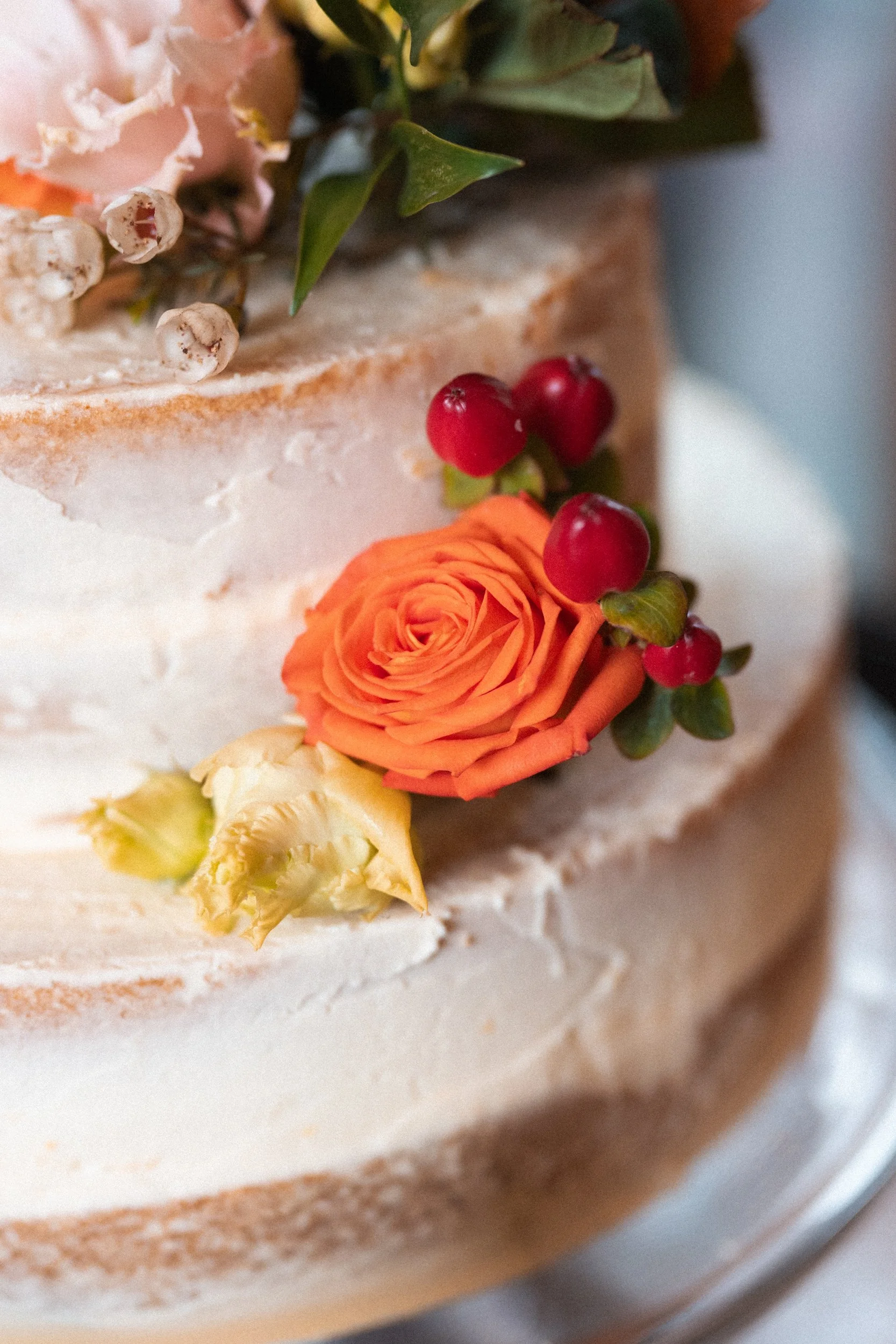 Close-up of a layered wedding cake decorated with an orange rose, a yellow rose, and red berries, with a white frosting finish.