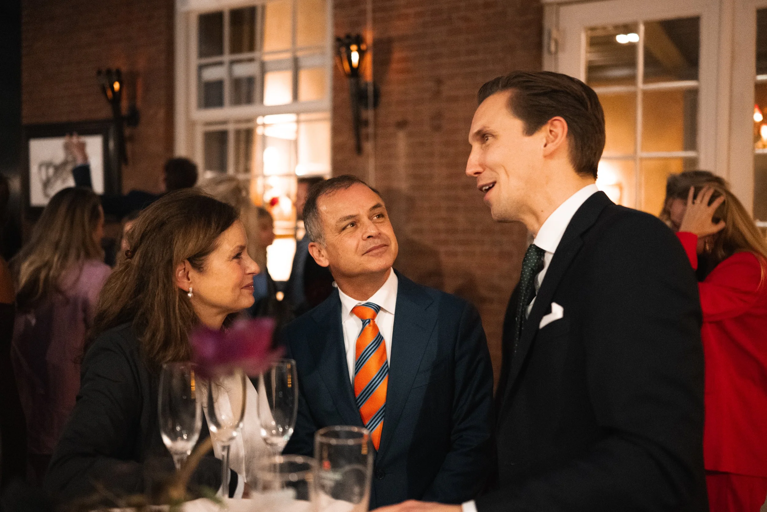 Three people engaged in conversation at a formal event or party, with glasses on a table in the foreground and other guests in the background.