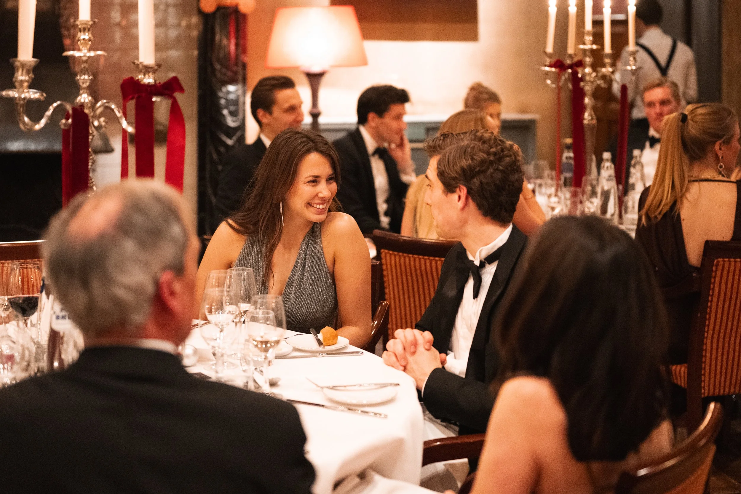 A formal dinner with guests in elegant attire, smiling and engaging in conversation at a restaurant or banquet hall decorated with candles and warm lighting.