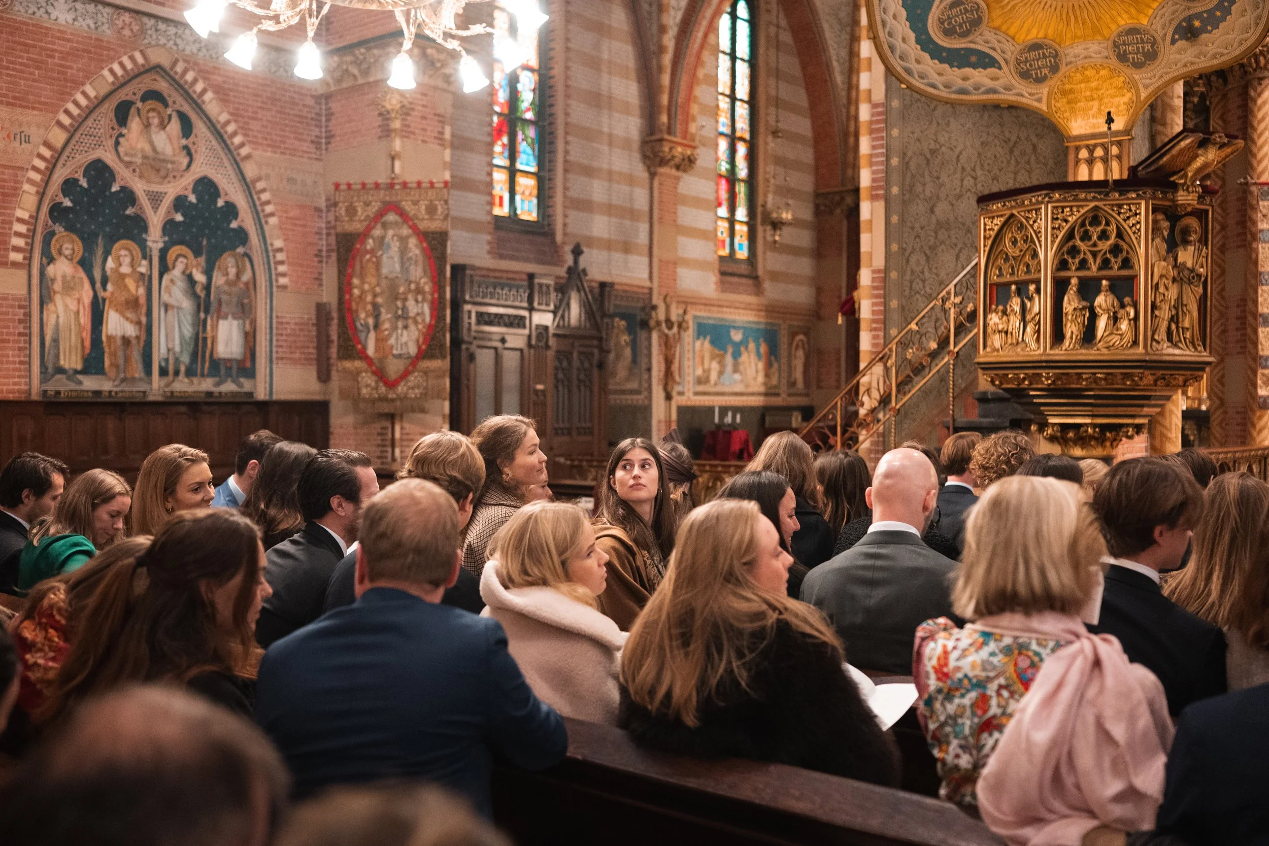 People seated in a church during a service or event, with stained glass windows and ornate religious artwork and decorations visible in the background.