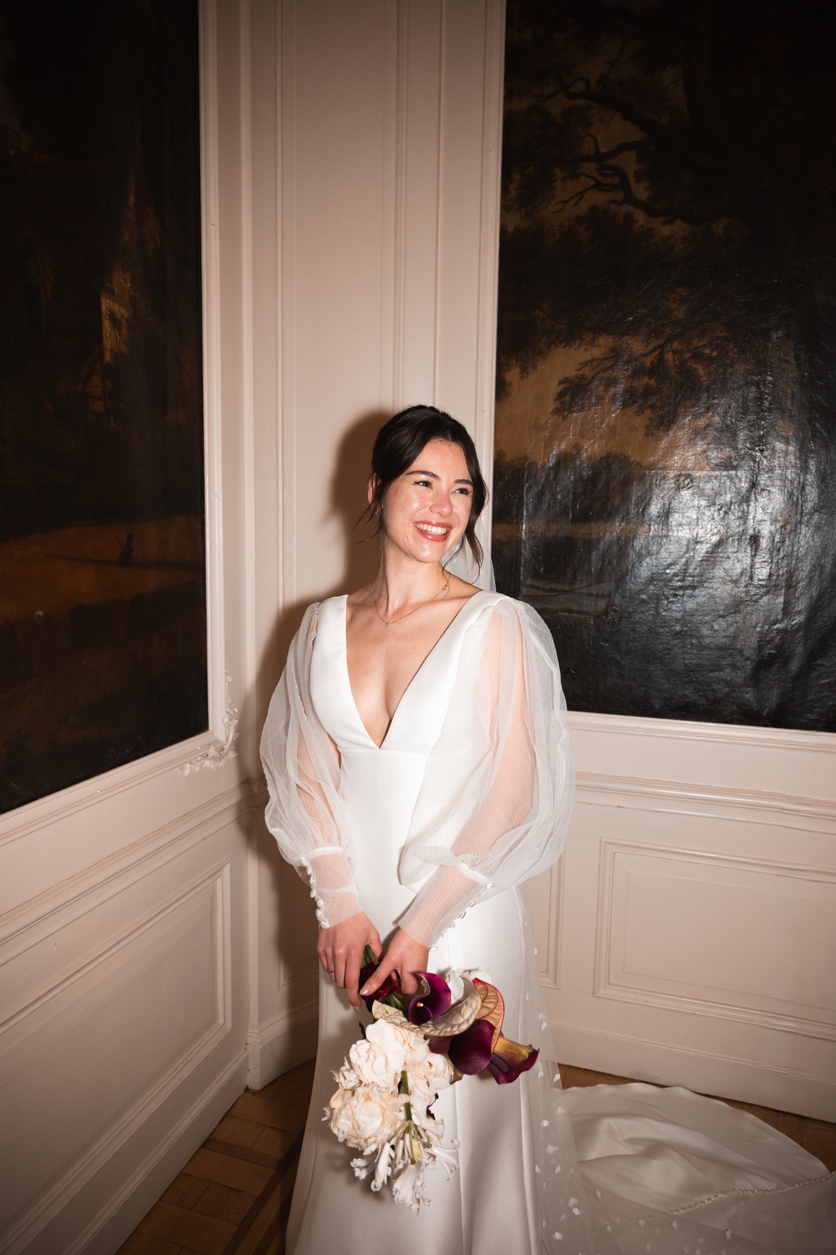 A woman in a white wedding gown holding a bouquet, smiling in an elegant room.