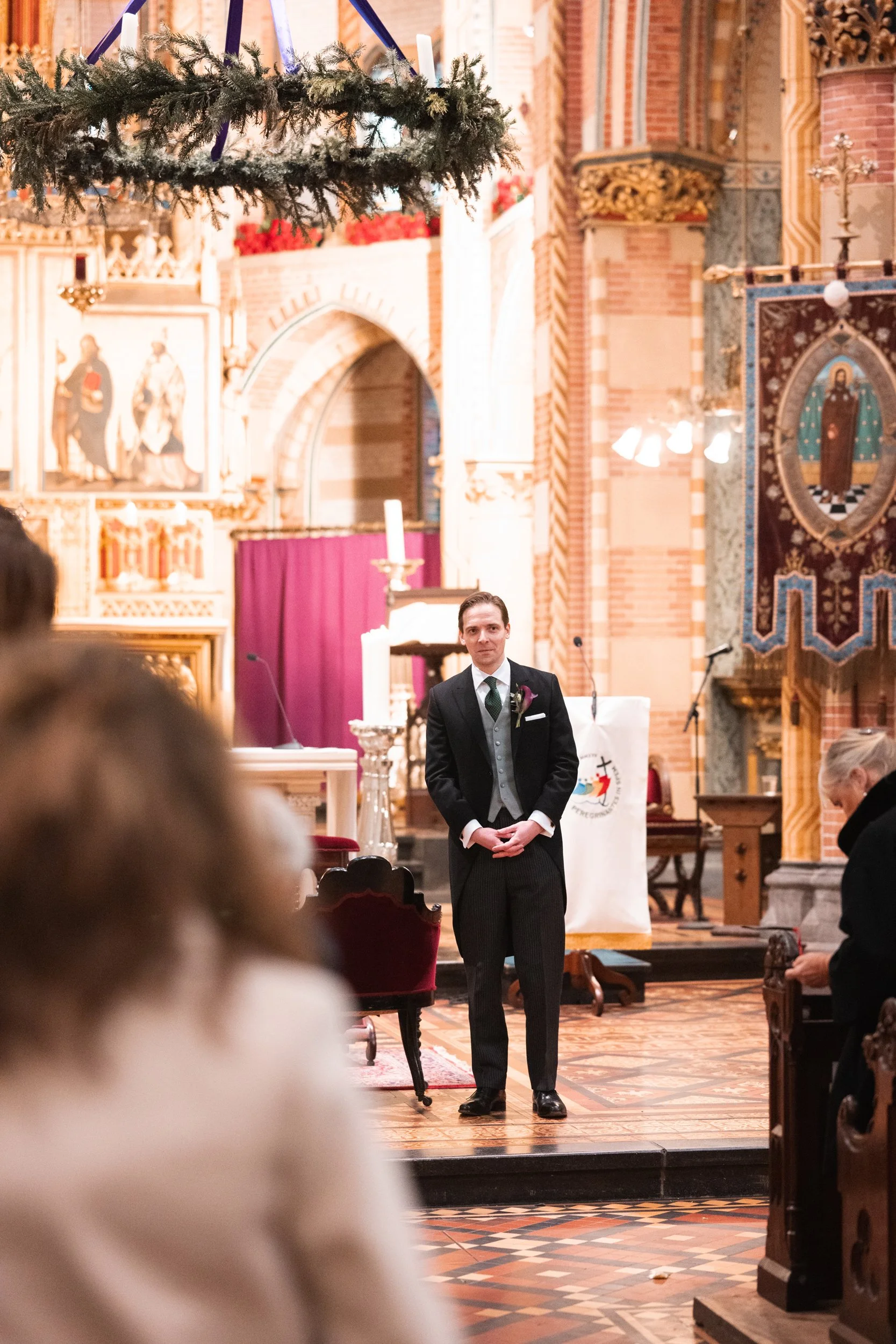 A groom in a black tuxedo and vest standing at the altar inside a church decorated for a wedding, with guests seated and a wedding officiant to the right.