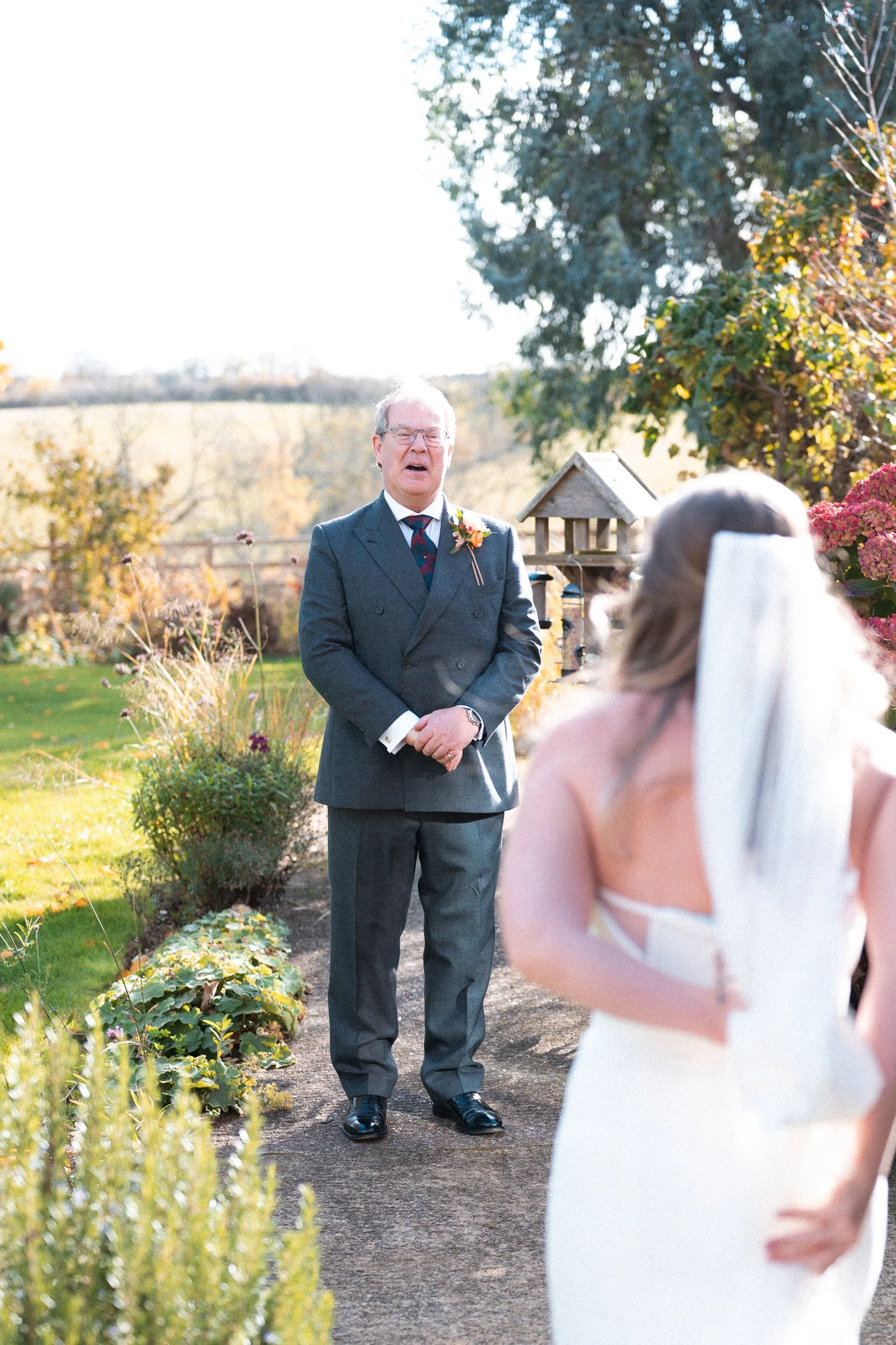 An outdoor wedding scene where an officiant, dressed in a gray suit, is singing or speaking while facing the bride. The bride, in a white wedding dress and veil, is standing with her back to the camera. The setting is a garden with trees, plants, and