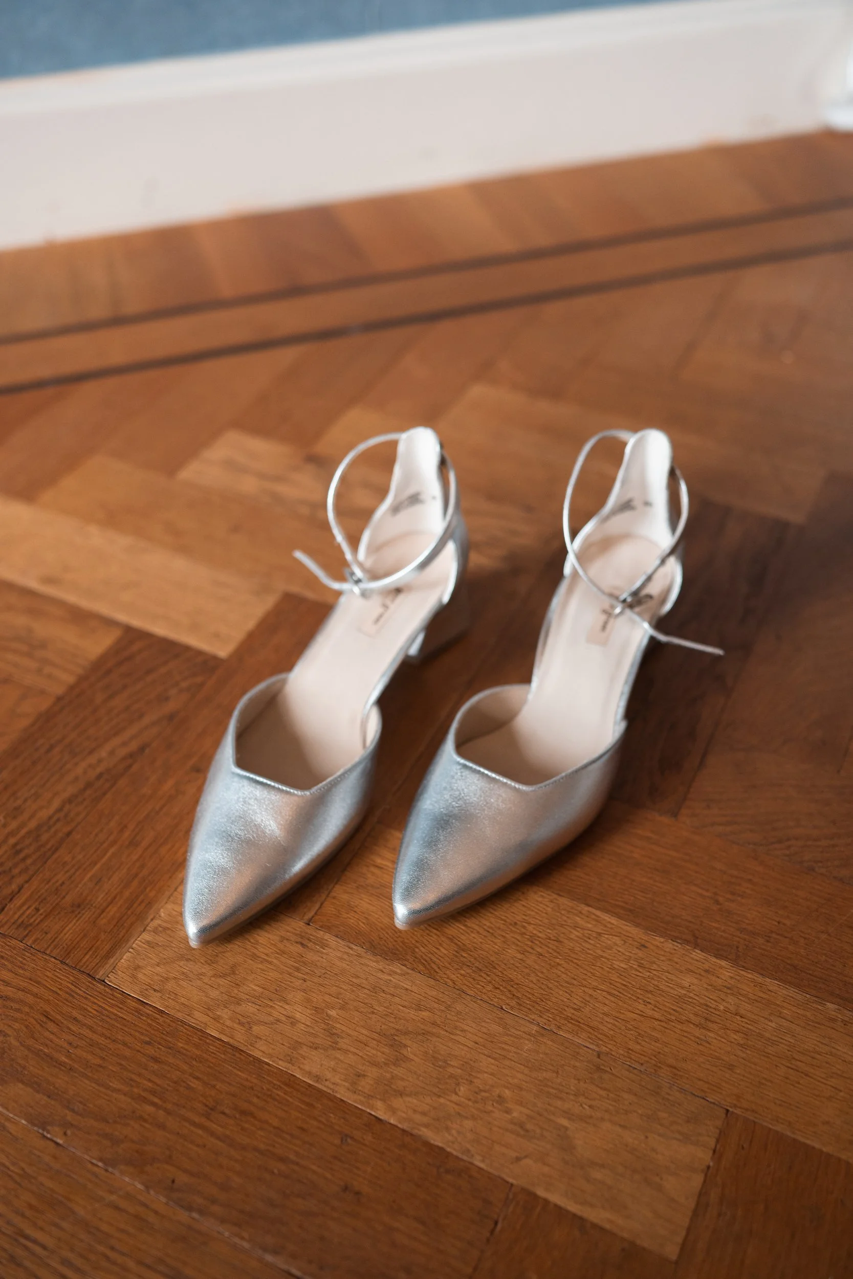 Silver high-heeled shoes with ankle straps placed on a wooden floor.