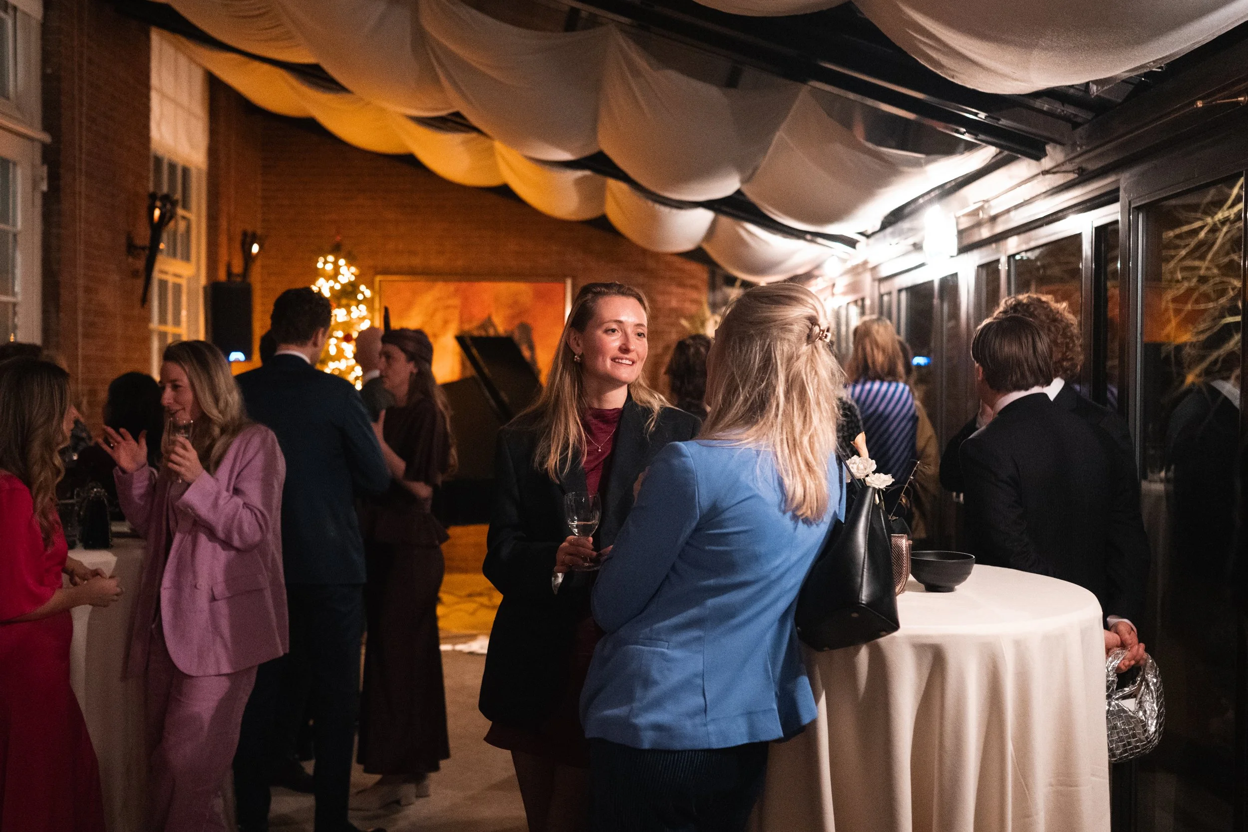 People socializing at a holiday party in an indoor venue with a brick wall, white fabric ceiling decorations, and a Christmas tree with lights in the background. Attendees are standing around high tables, holding drinks, and engaging in conversations