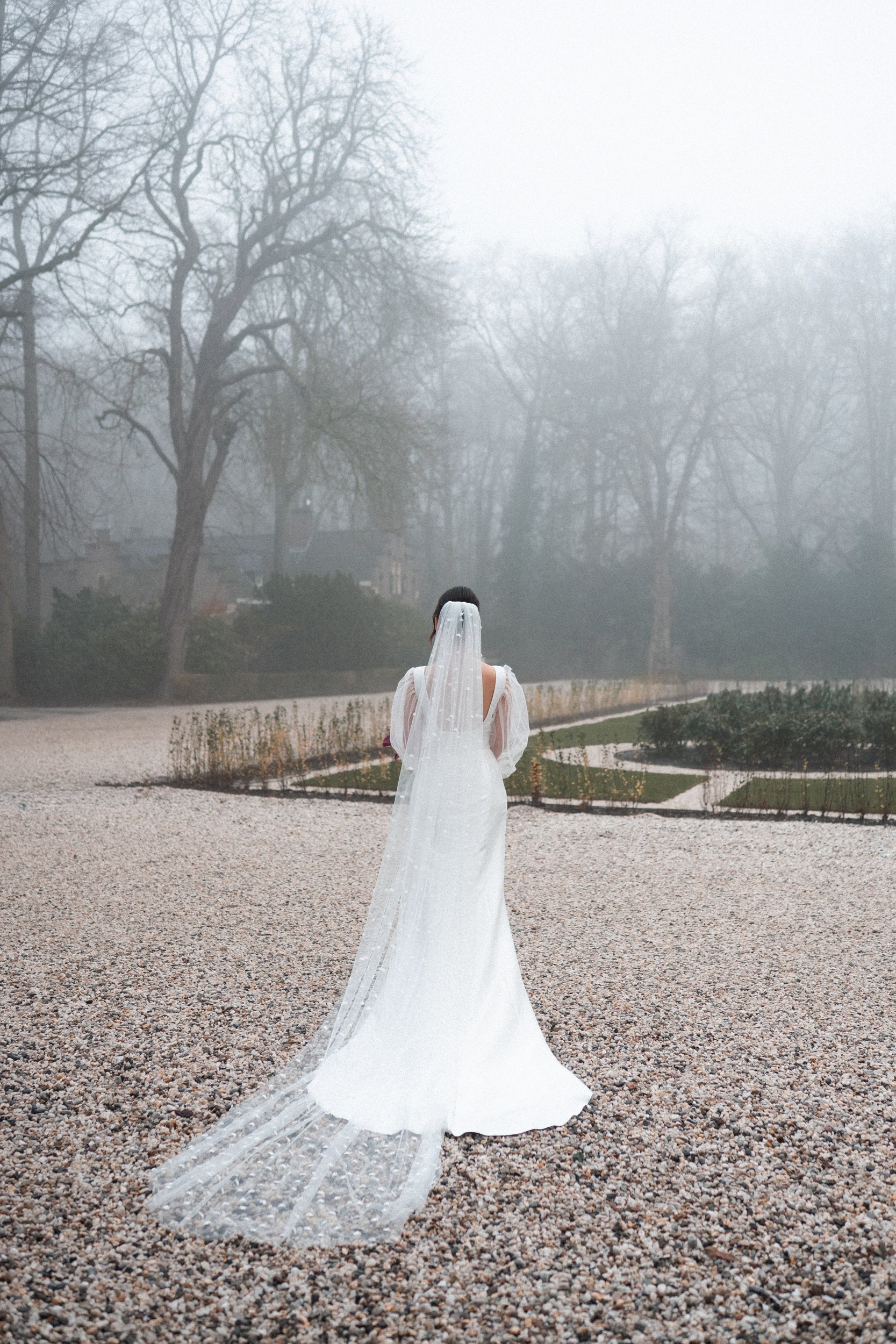 A bride in a white wedding dress and veil walking away on a gravel path in a foggy outdoor setting with leafless trees and garden beds.