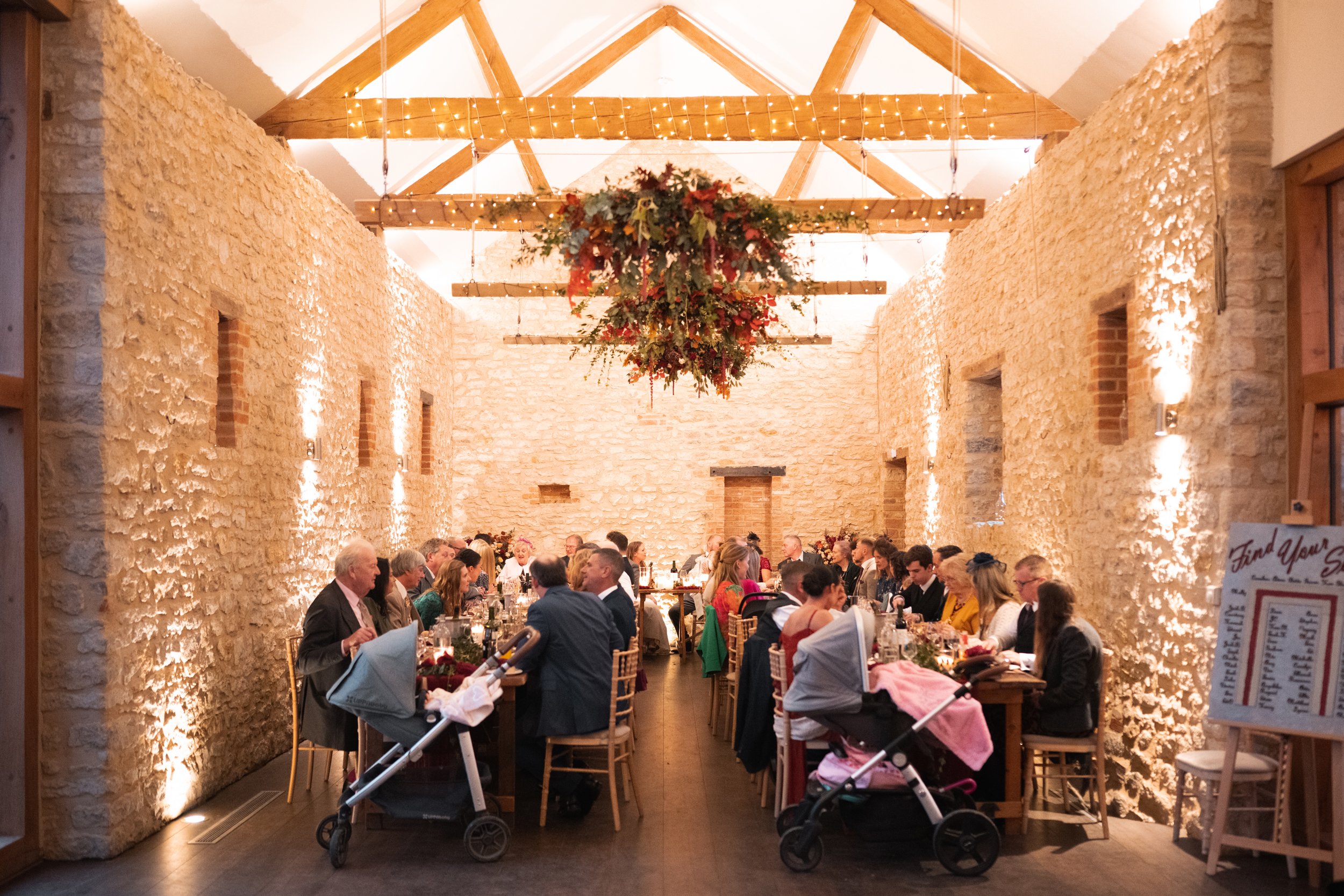 A large indoor wedding reception with stone walls, wooden beams with string lights, and a central floral chandelier. Guests are seated along a long table