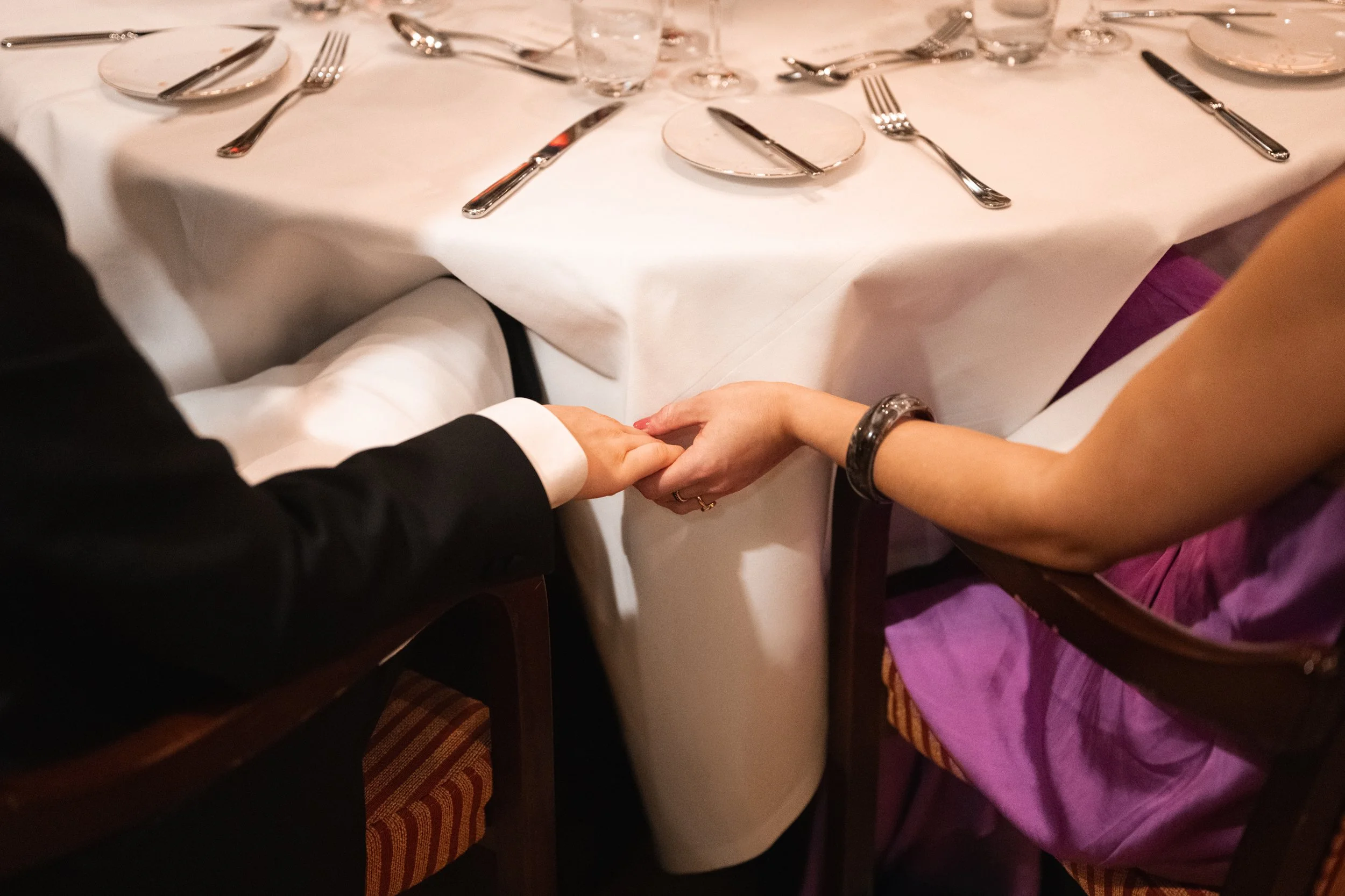 A couple holding hands underneath a white tablecloth at a formal dining table with silverware and glasses.