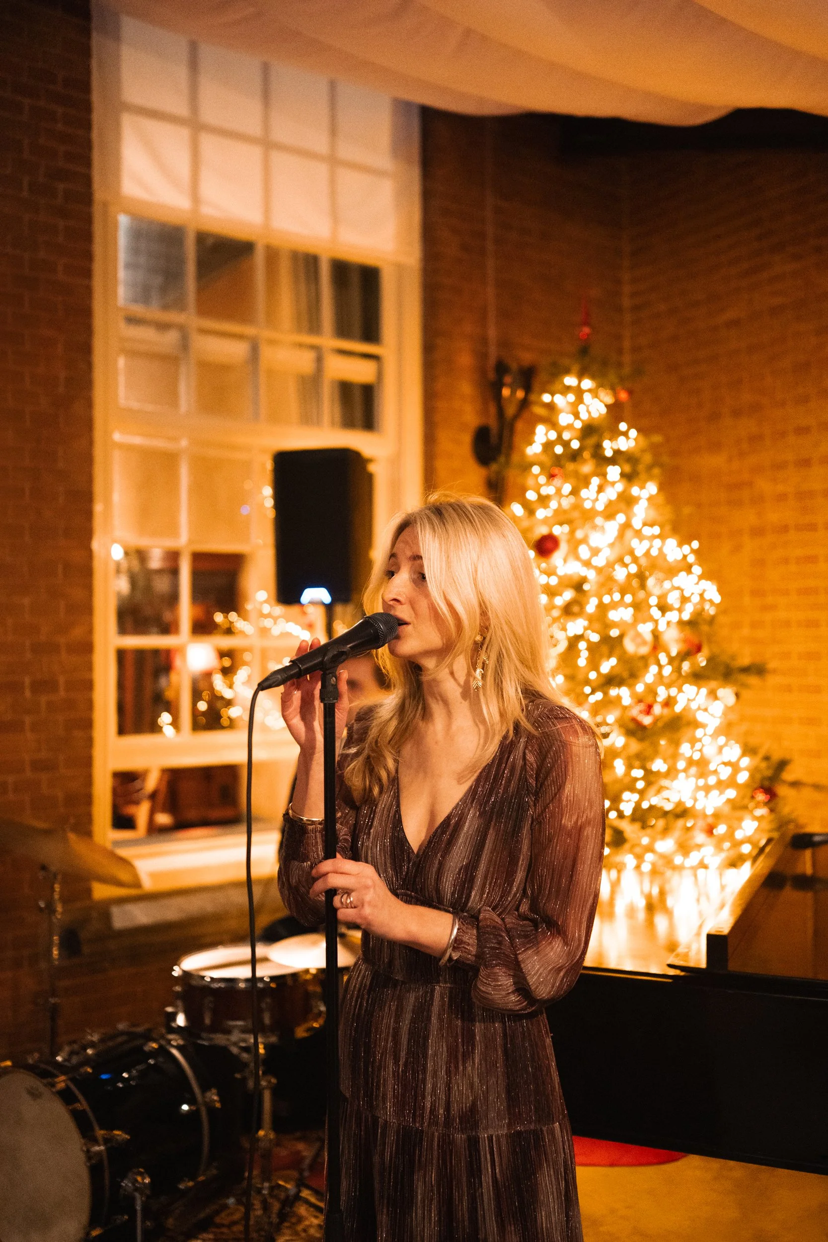 A woman singing into a microphone during a Christmas celebration, with a decorated Christmas tree with lights and ornaments in the background.