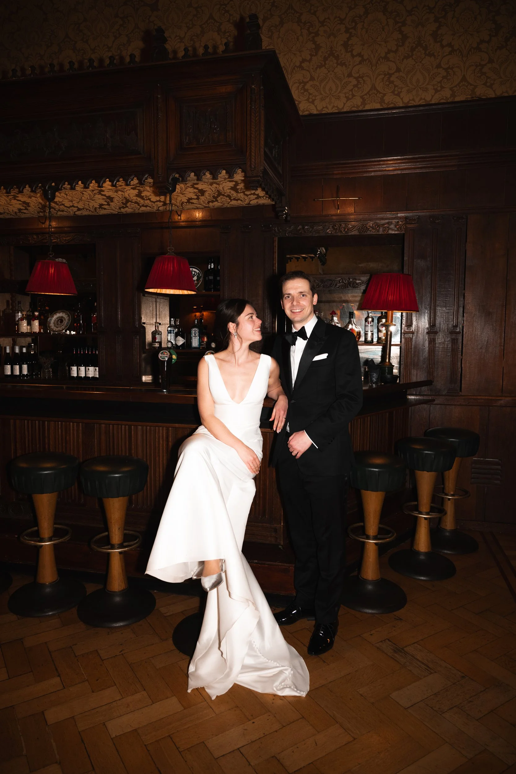 A bride and groom in wedding attire at a bar with dark wood paneling, red lampshades, and black chairs.