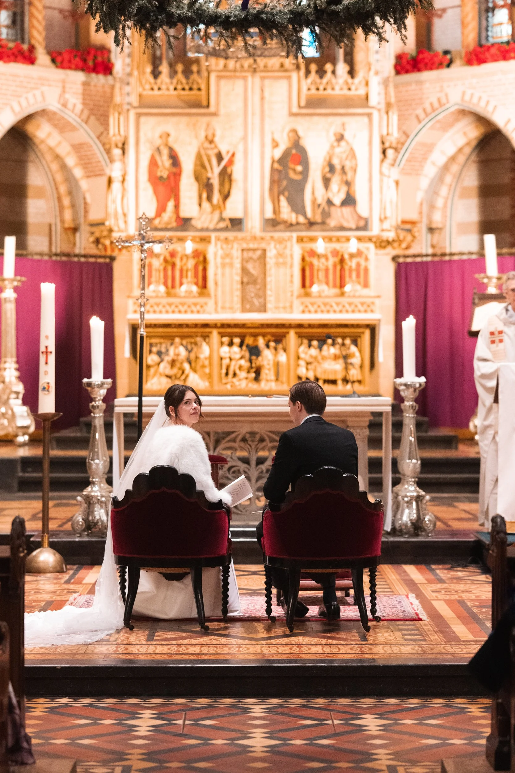 Bride and groom seated in church during wedding ceremony, facing the altar with large religious paintings and candles.