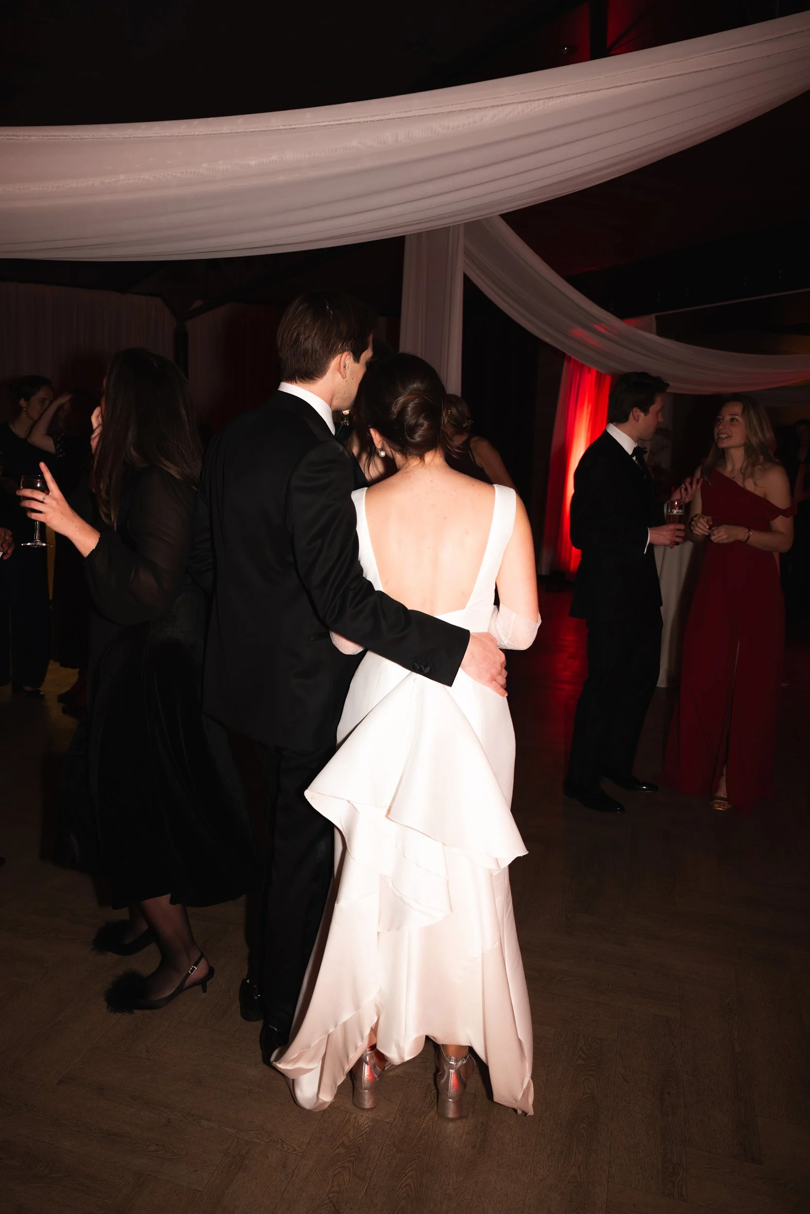 Couple dancing at a formal event with others socializing in the background, decorated with draped fabric and dim lighting.