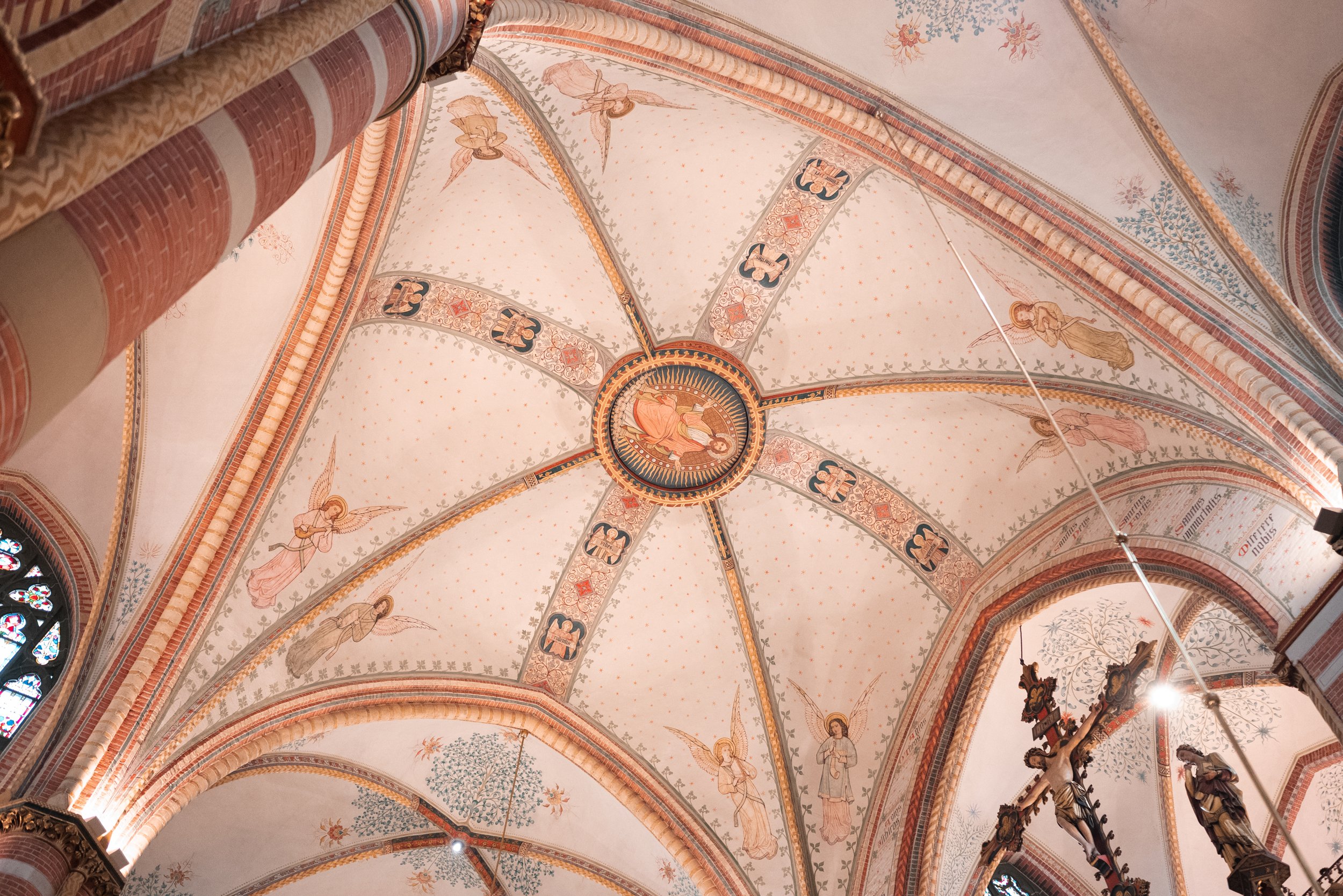 Ceiling of a church with religious artwork, including angels and a central circular icon of Jesus Christ, with stained glass windows and ornate decorations.
