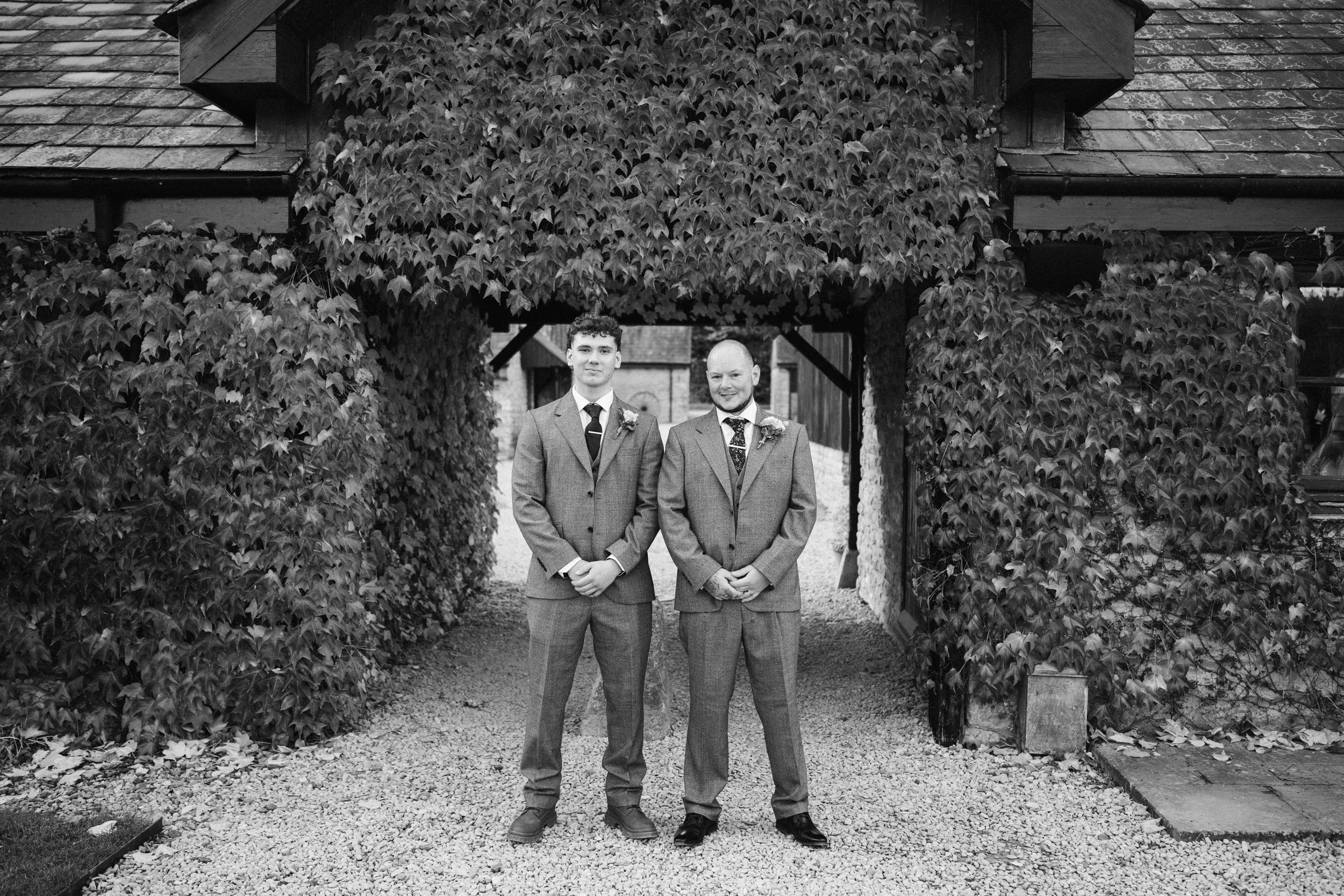 Two men in suits standing side by side outdoors in front of a leafy archway and stone building, posing for a photo.