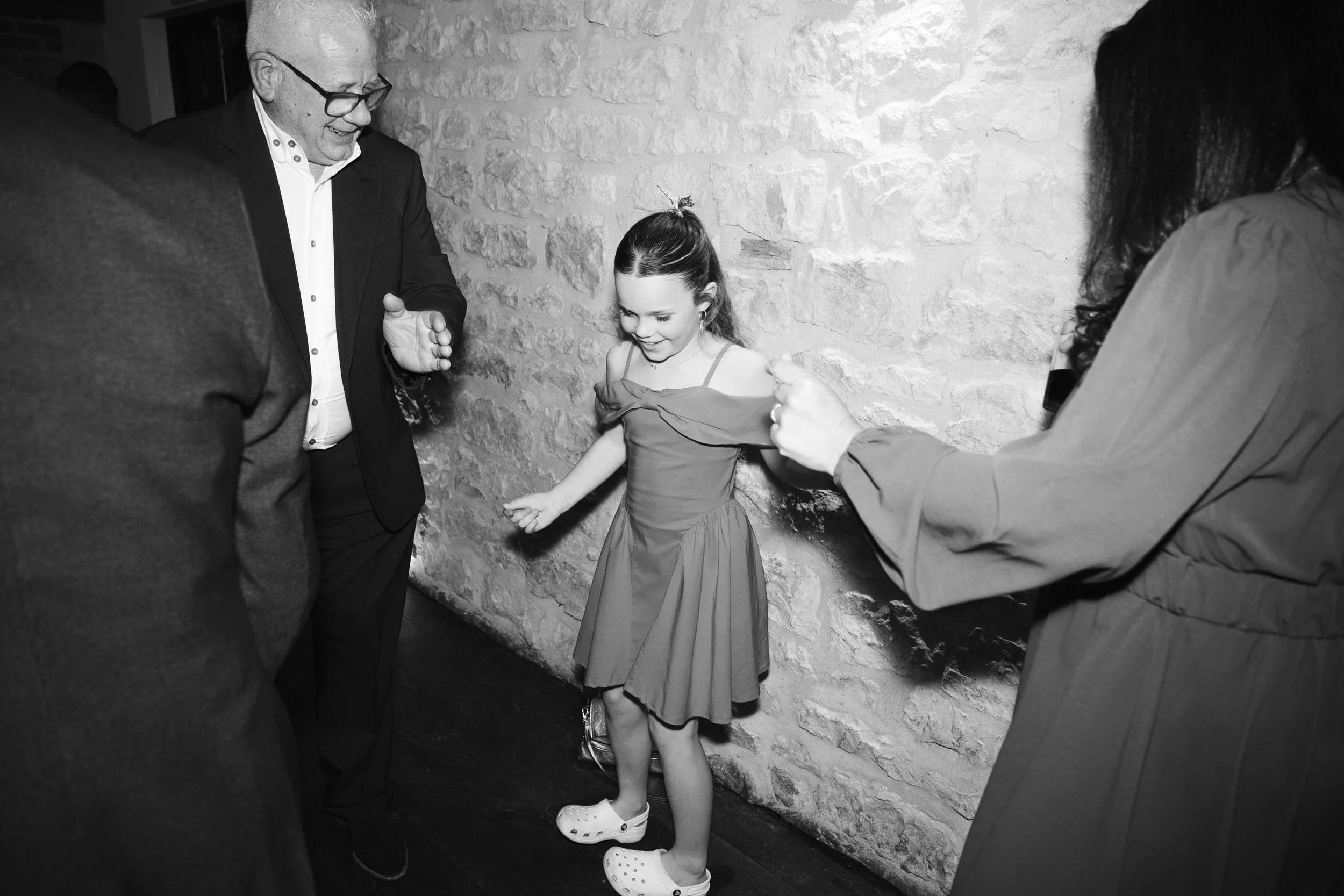 A girl in a dress with crutches standing against a brick wall, smiling, with three adults around her, one with glasses and a suit, and two women, one with long dark hair, all engaging and smiling at her.