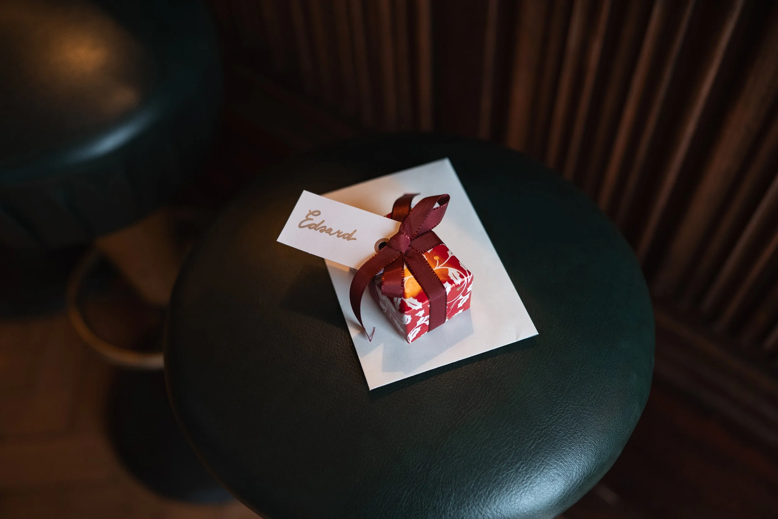 Small gift box with red floral pattern, tied with a maroon ribbon, placed on a white paper on a dark green cushioned stool.