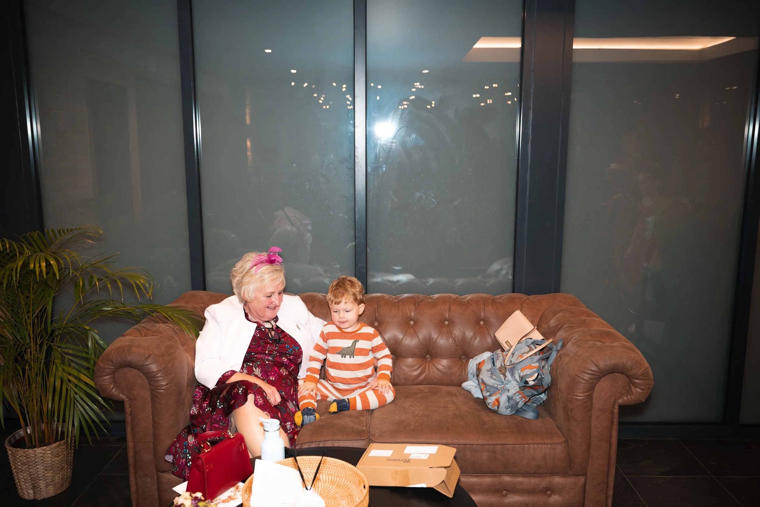 An elderly woman and a young boy sitting on a brown leather couch in a modern room, with a large plant to the left and a glass wall behind them.