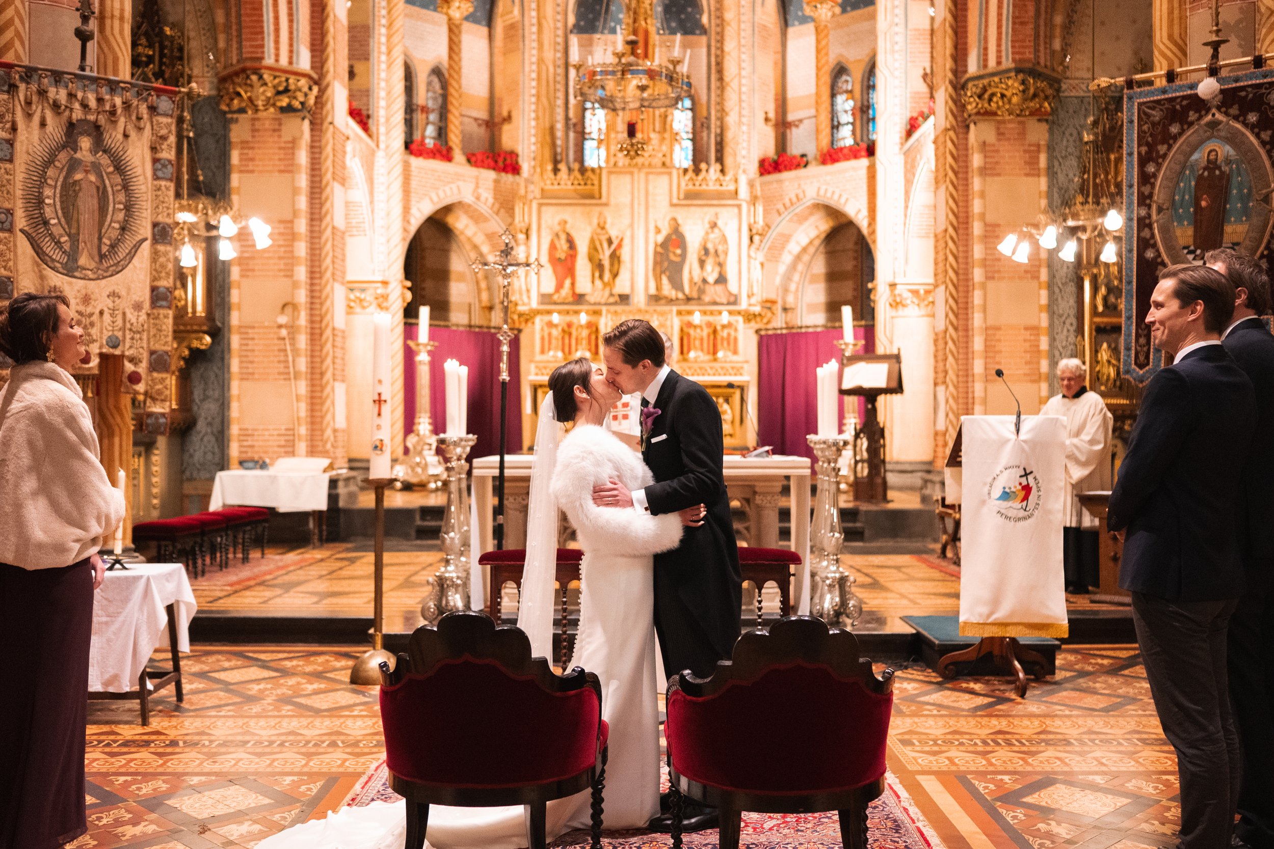 A couple getting married in a church, kissing at the altar, with a priest officiating. The church is ornate with candles, religious artwork, and red curtains. Guests and witnesses are standing nearby.