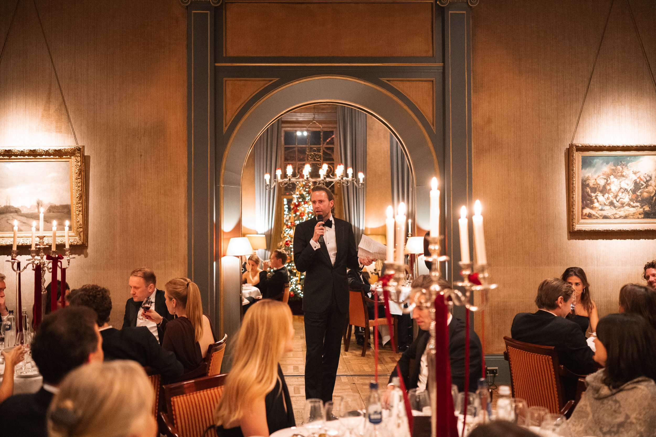 A man in a tuxedo giving a speech at a formal dinner event, with guests seated at tables, a Christmas tree, and festive decorations in a warmly lit elegant room.