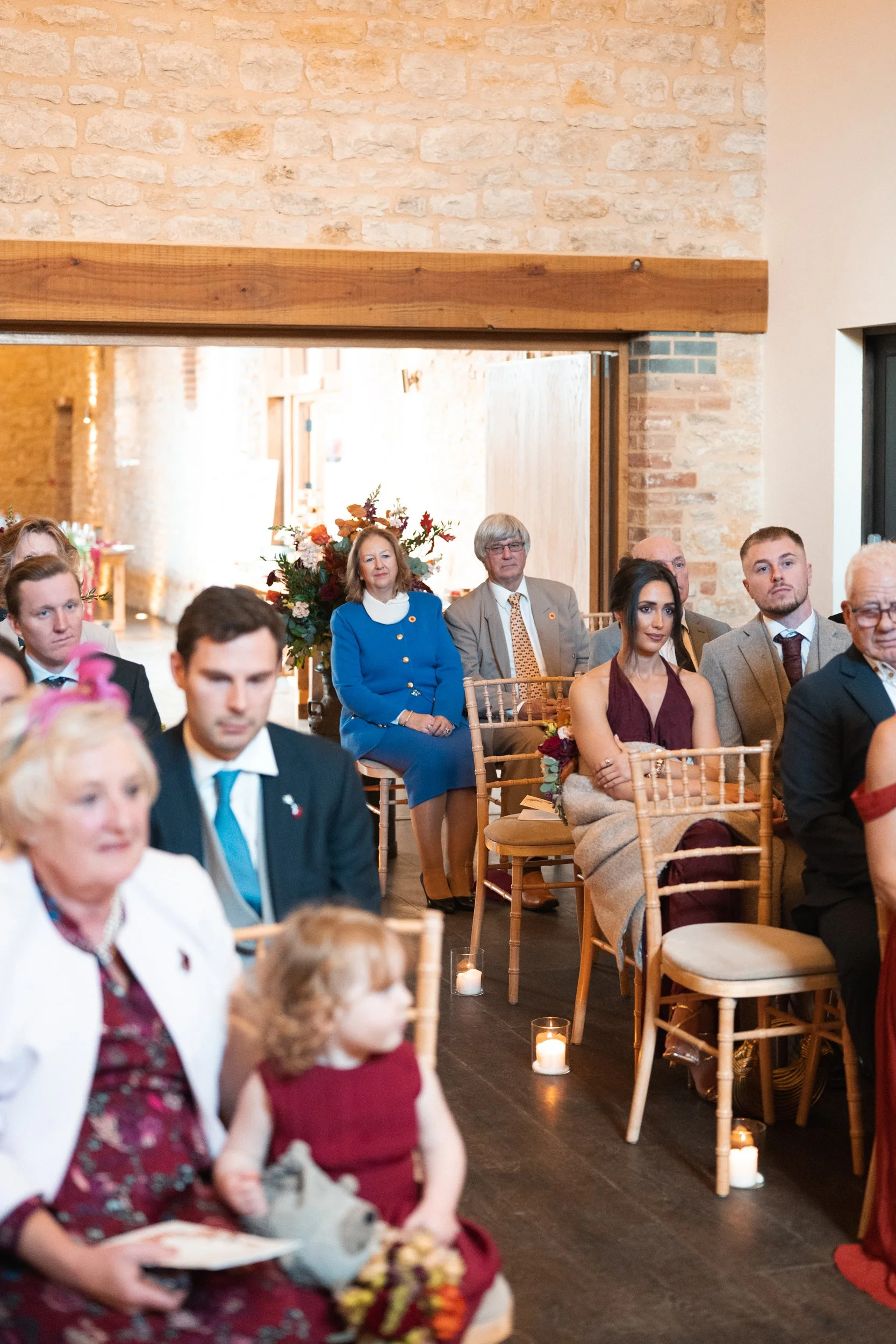 Attendees seated in a rustic venue with brick walls, candles on the floor, and floral arrangements, attending a formal event or wedding.