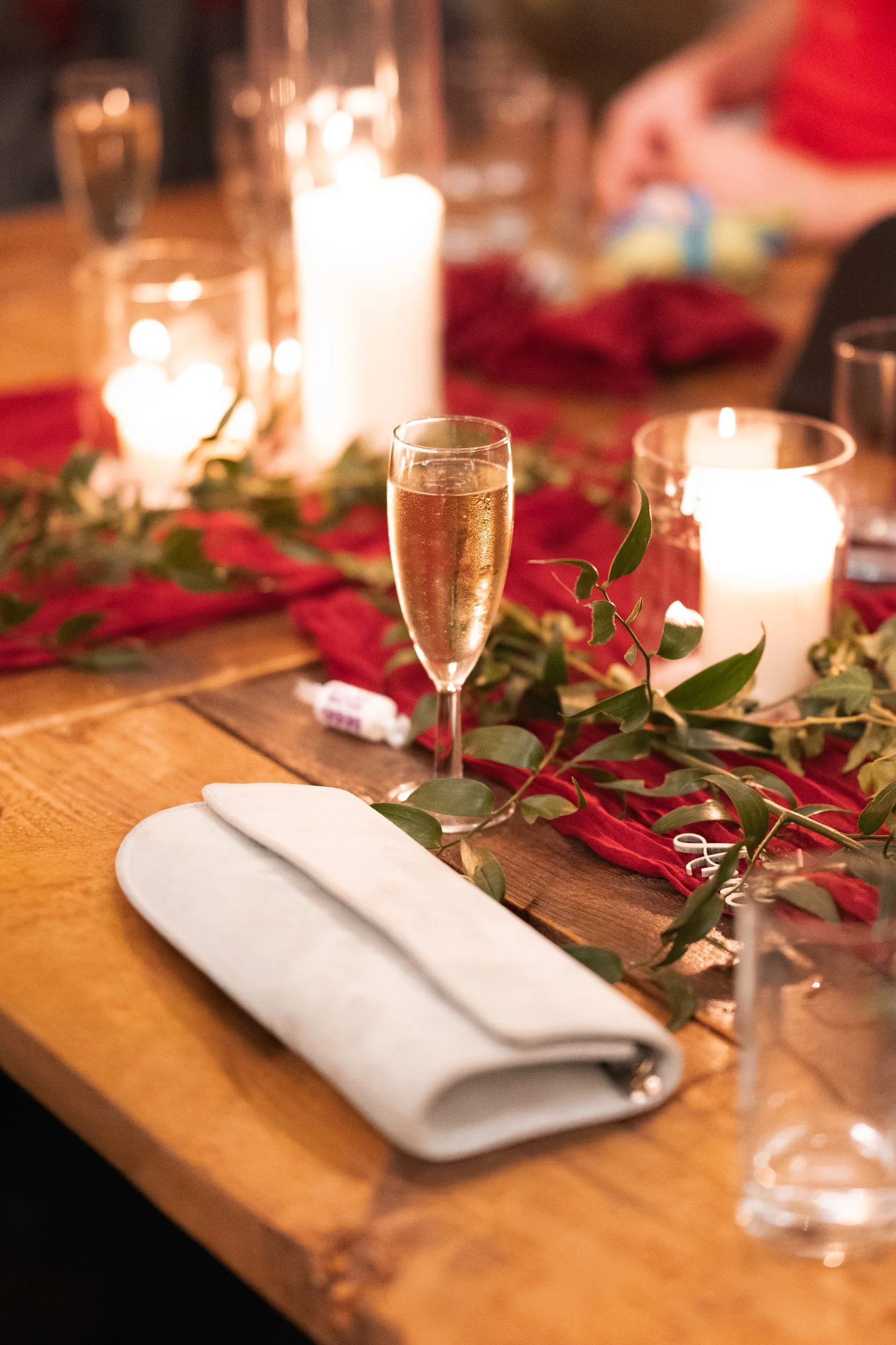 A wooden table set for a celebration with a champagne flute, white napkin, and candles with greenery and red fabric decoration.
