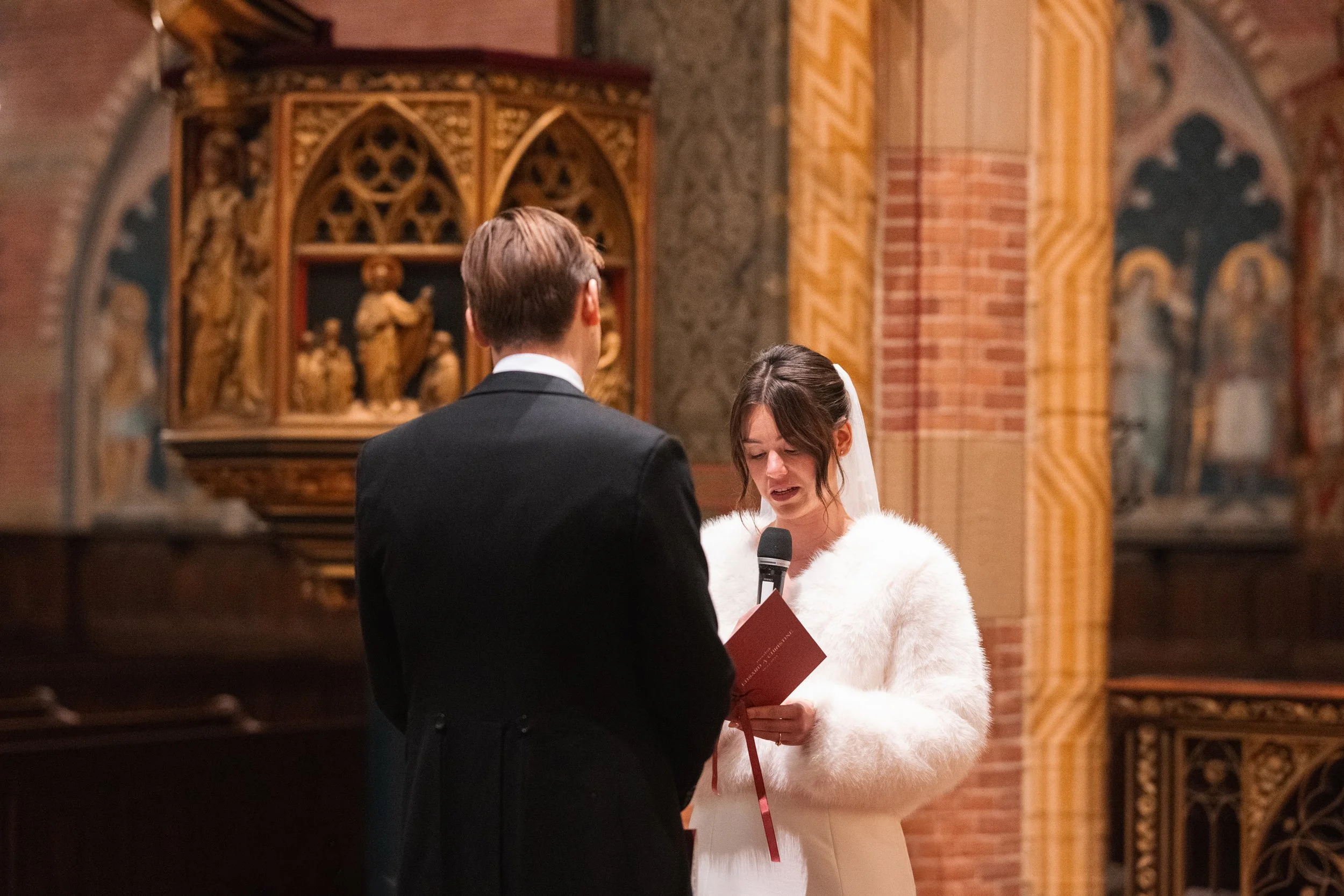 A bride and groom exchanging vows during a wedding ceremony inside a church. The bride is reading from a booklet and holding a microphone, wearing a white fur coat and veil. The groom is dressed in a black suit, facing the bride with his back to the 