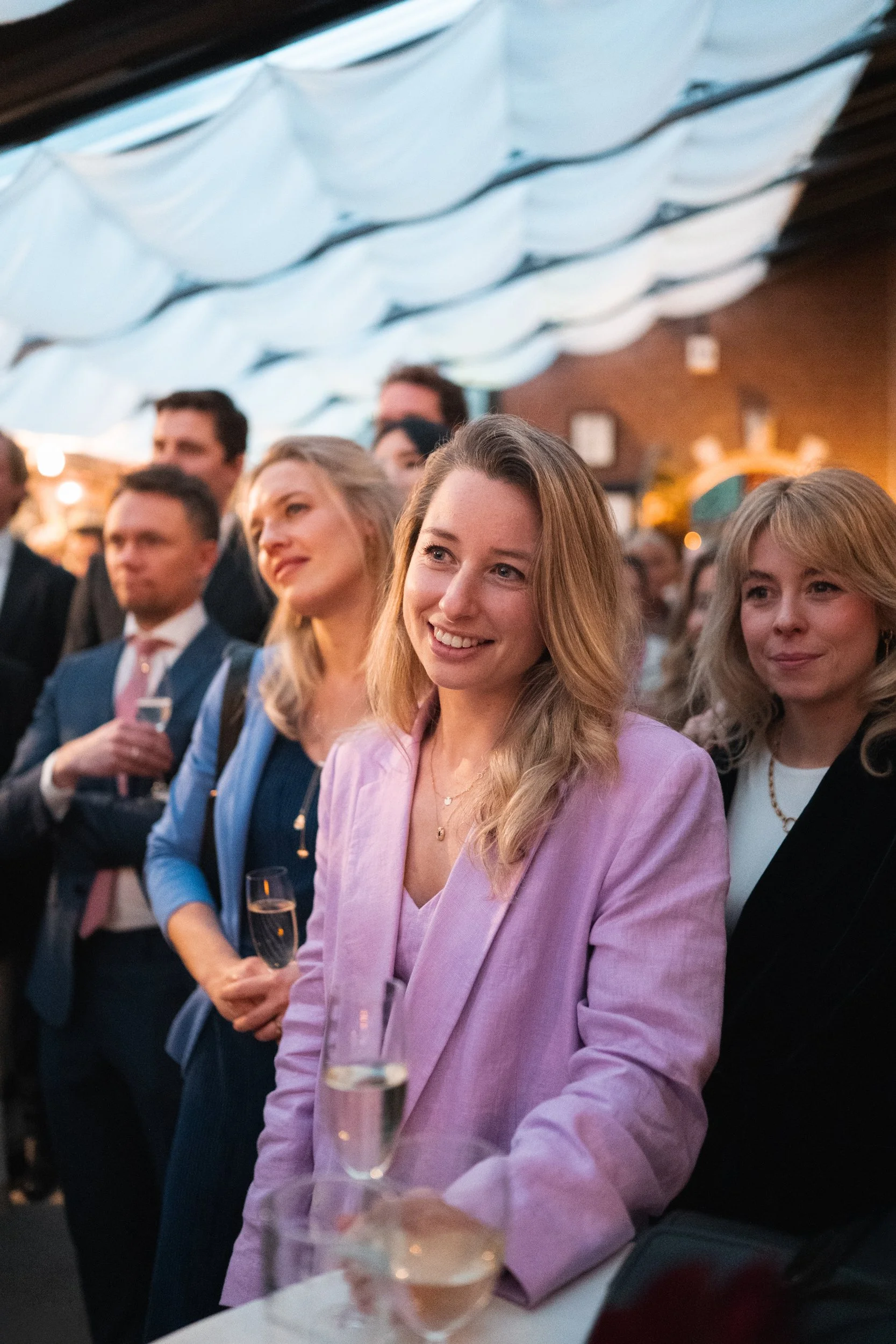 A group of well-dressed people at a social event or celebration, with a woman in a pink blazer smiling and holding a glass of champagne in the foreground.