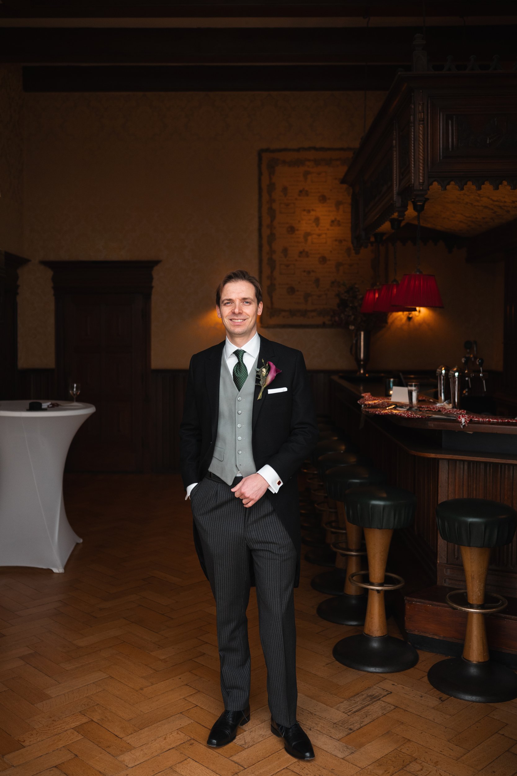 A man dressed in formal wedding attire standing in a dimly lit bar with wood paneling and bar stools.
