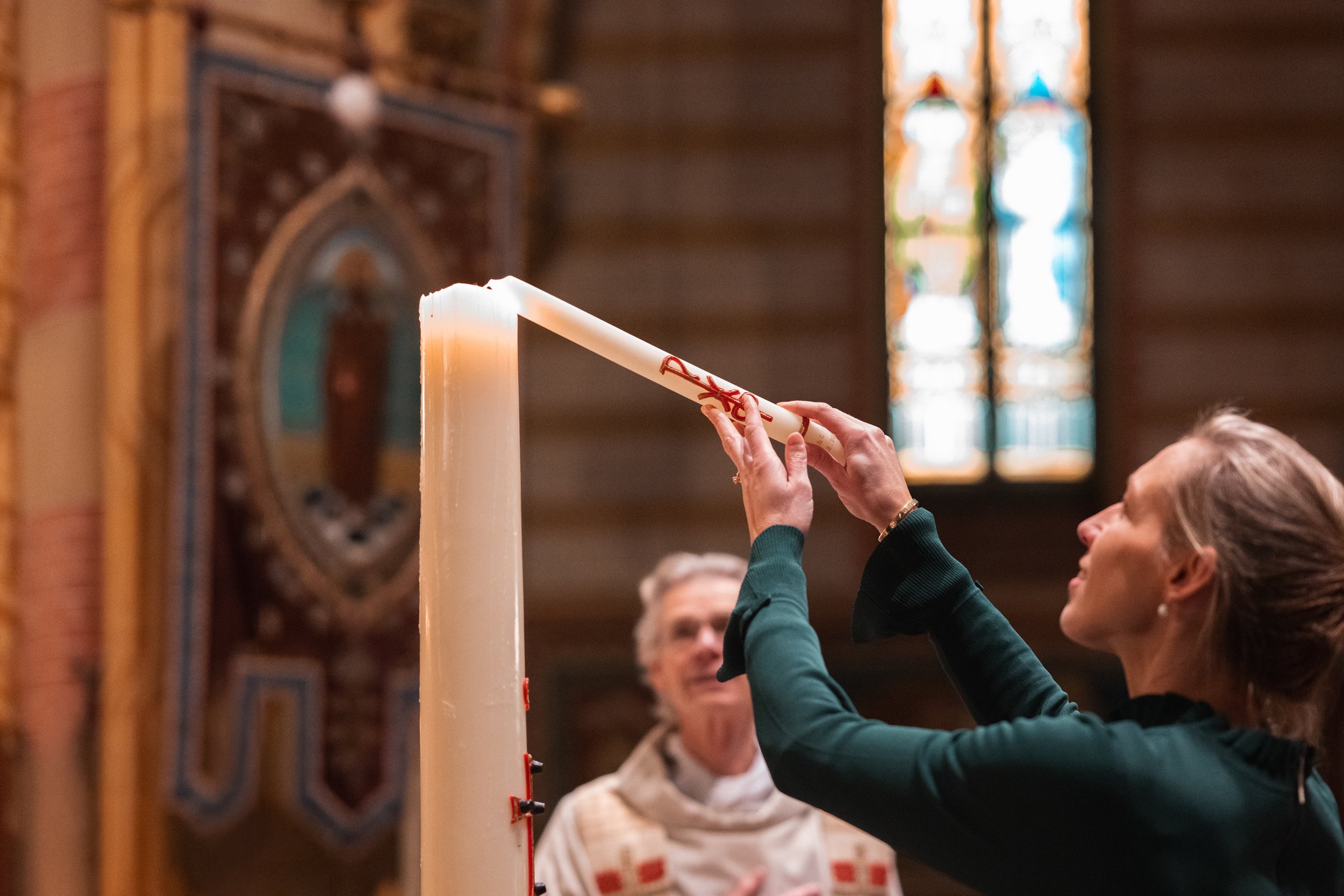 A woman lights a large candle in a church, with a priest looking on in the background.