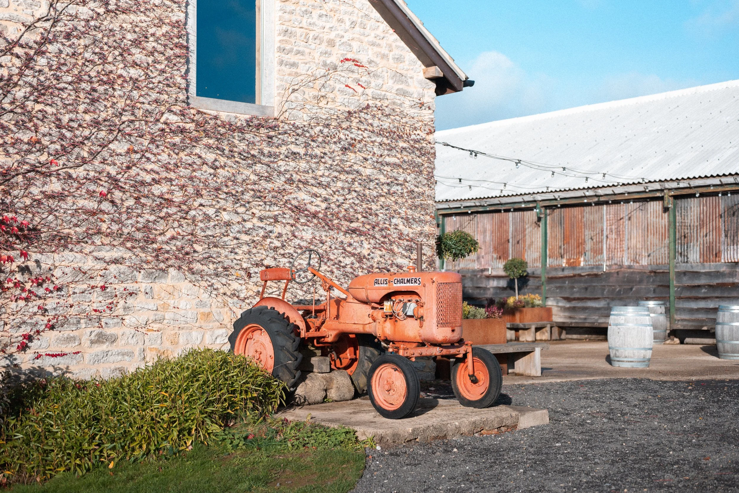 An orange vintage tractor is parked outside next to a stone wall with some pink flowering vines. The background shows a building with a corrugated metal roof, some potted plants, and a wooden fence, under a clear blue sky.