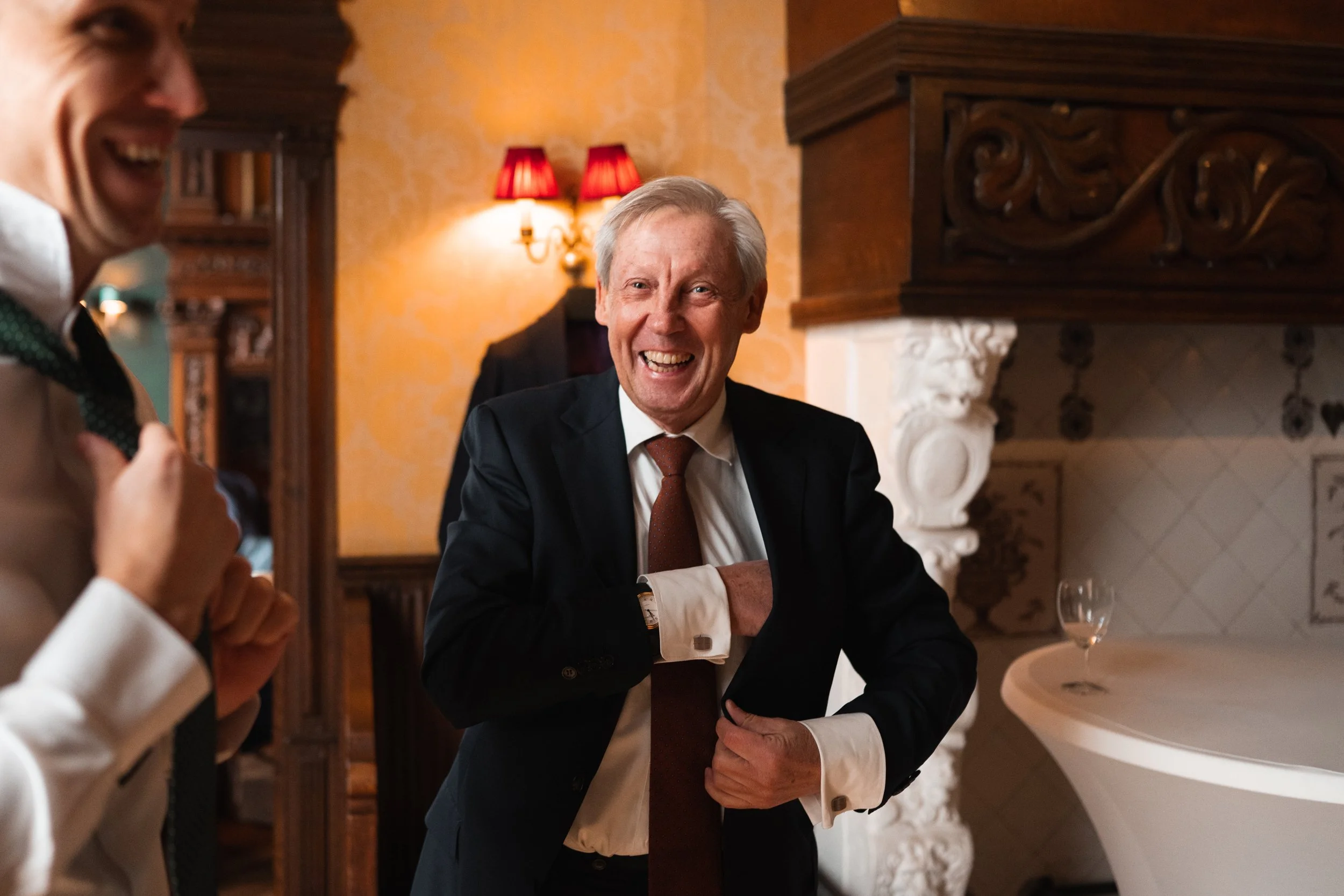 Two men in formal attire are laughing and smiling in a warmly decorated room with ornate woodwork and a fireplace. One man is adjusting his suit jacket, and the other is visible on the left side of the image.
