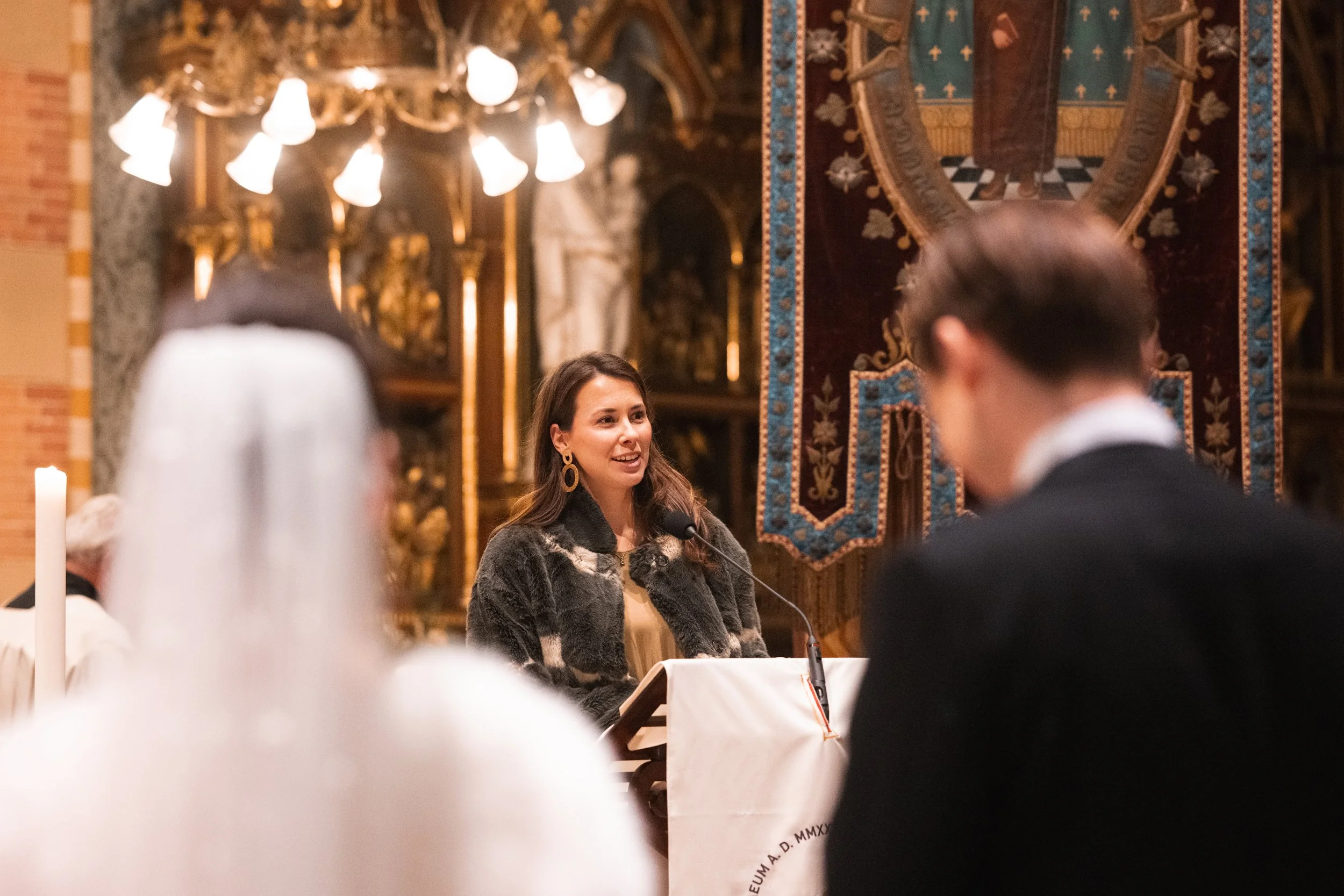 A woman speaking at a podium in a church or cathedral setting, with a journalist or speaker facing her in the foreground and an ornate religious banner behind her.