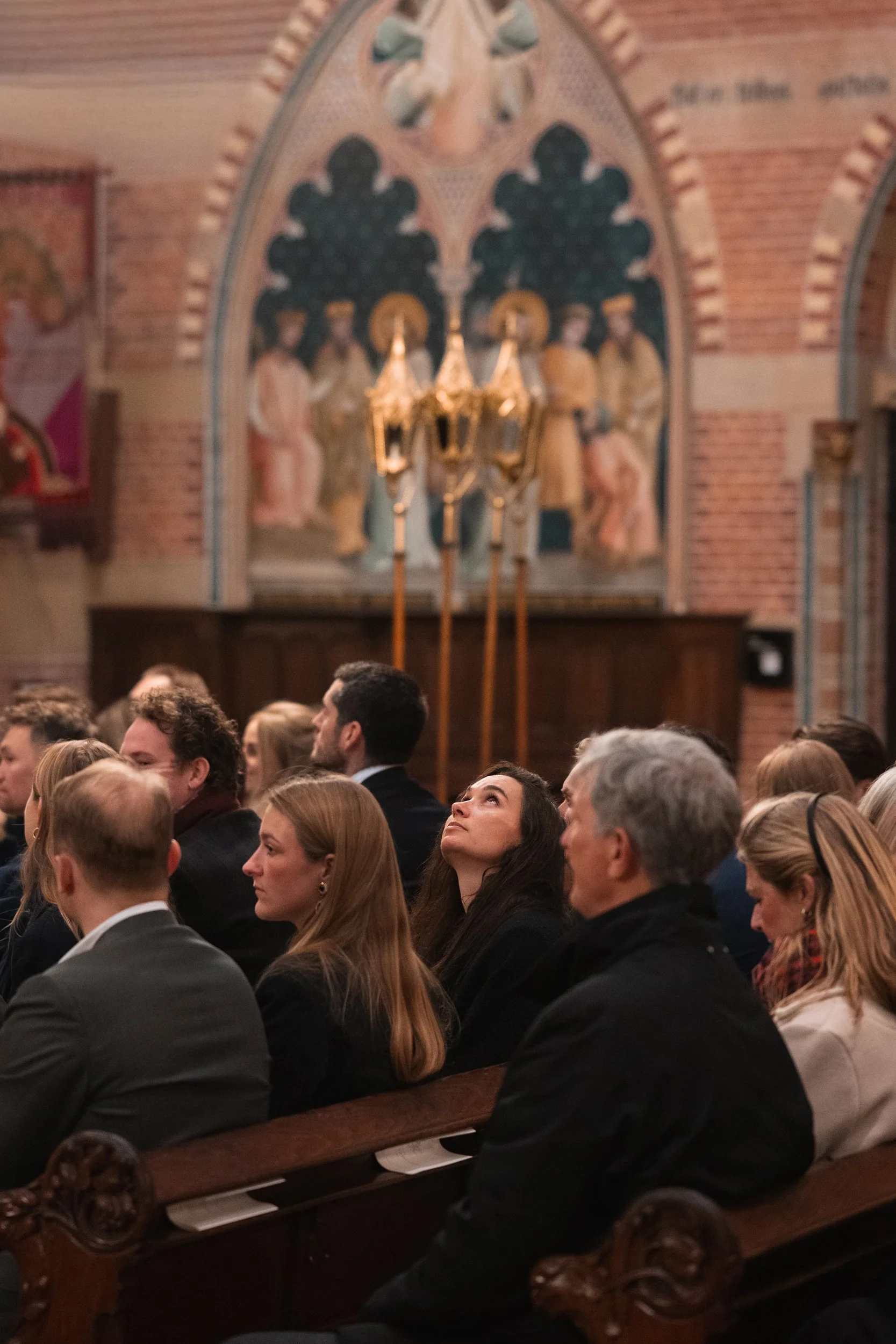 A group of people seated in a church or auditorium, paying attention to an event or speaker, with ornate religious artwork and a chandelier in the background.