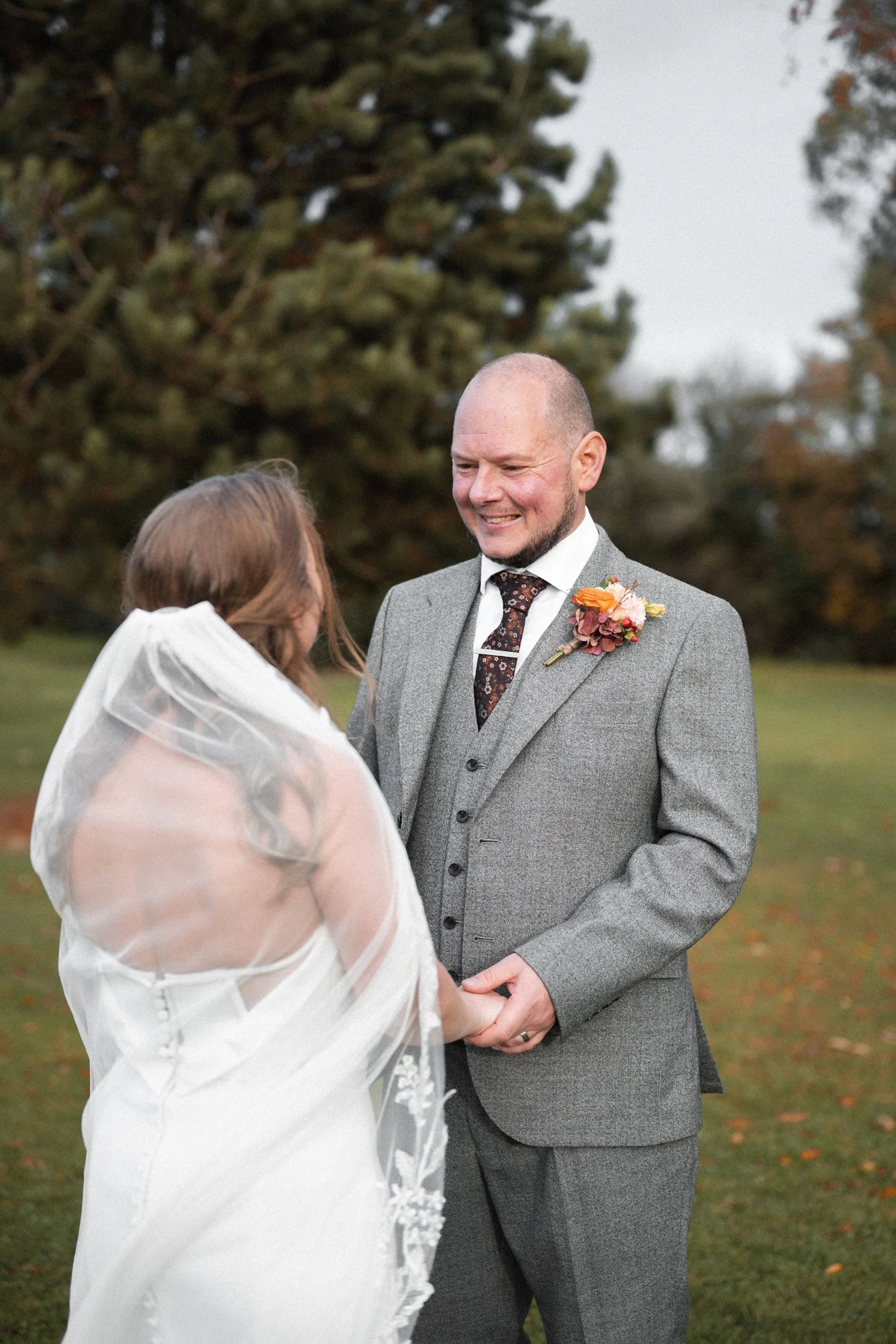 A bride and groom holding hands during their outdoor wedding ceremony on a cloudy day, with trees in the background.