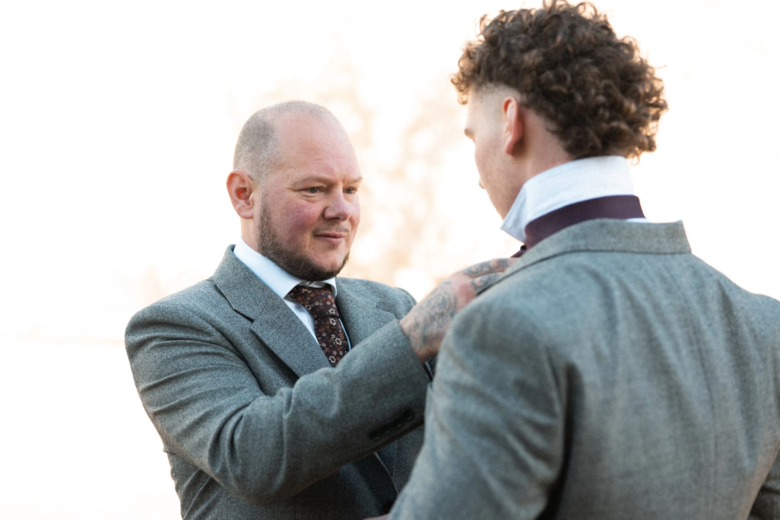 Two men in suits engaged in a handshake or greeting outdoors with a blurred background.