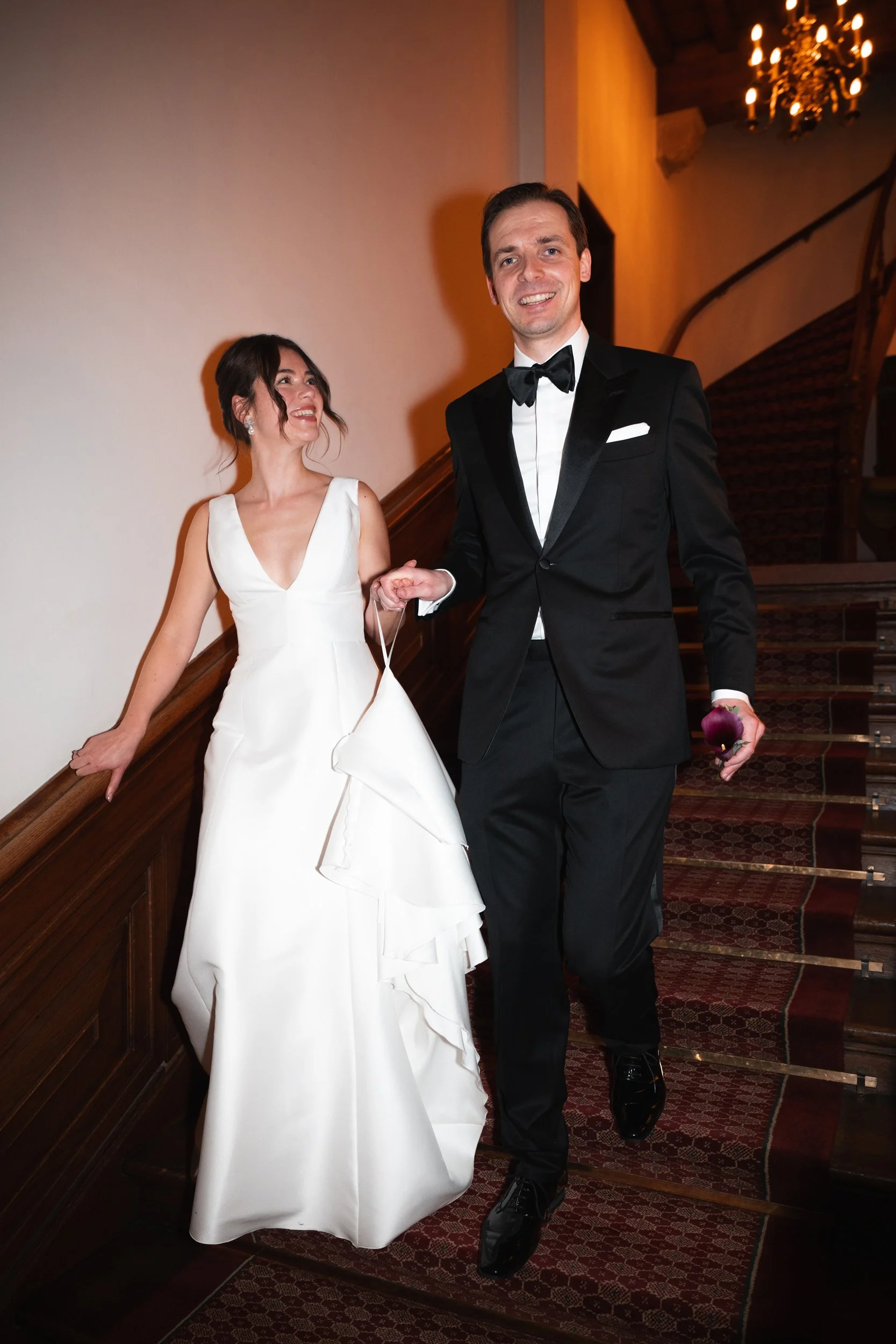 A bride and groom walking down a staircase at their wedding ceremony, holding hands, smiling, and dressed in formal wedding attire. The bride is wearing a white wedding gown, and the groom is wearing a black tuxedo with a bow tie.