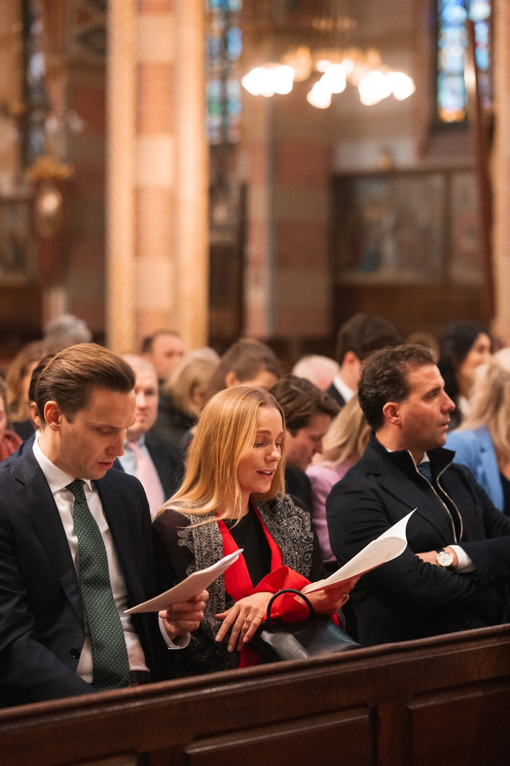 People attending a church service, seated in pews, reading programs, inside a church with stained glass windows and wooden decor.