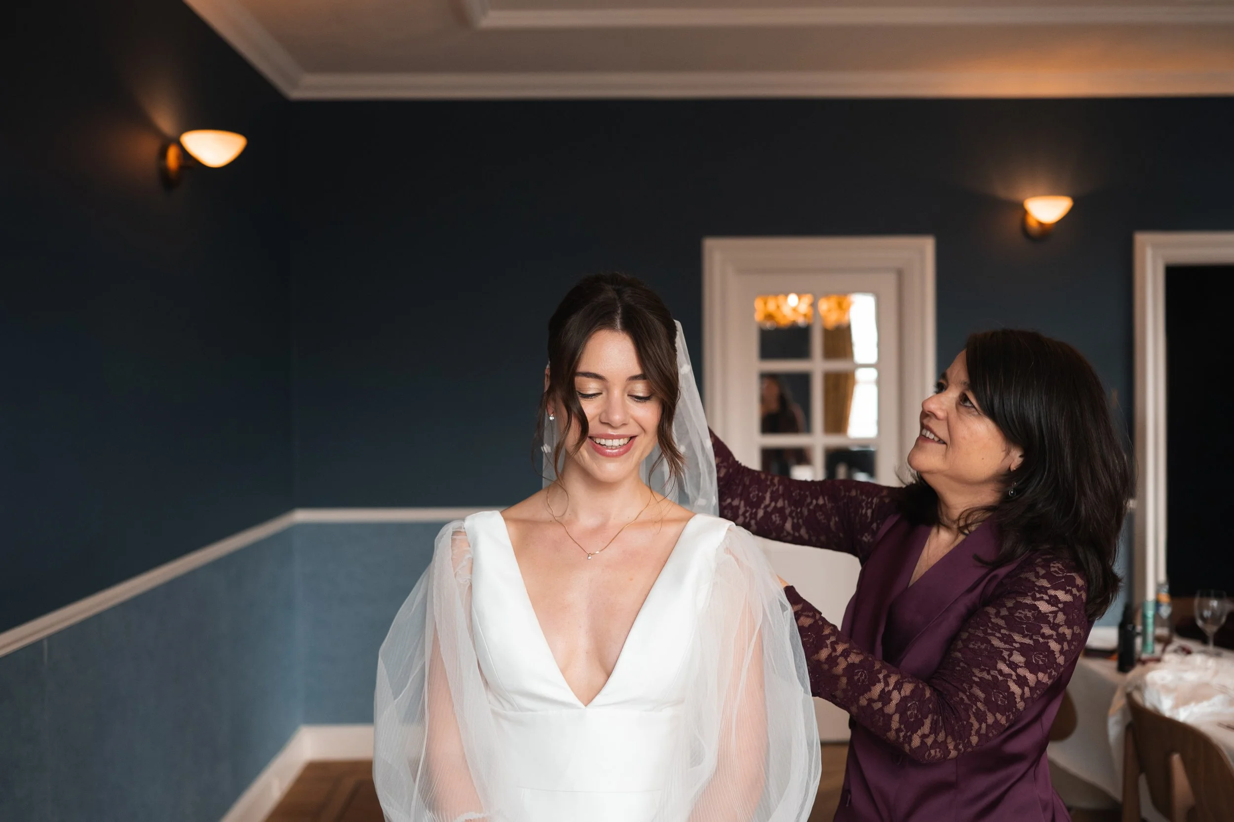 A bride in a white wedding dress with sheer sleeves smiling as a woman in a purple lace dress adjusts her veil in a warmly lit room.