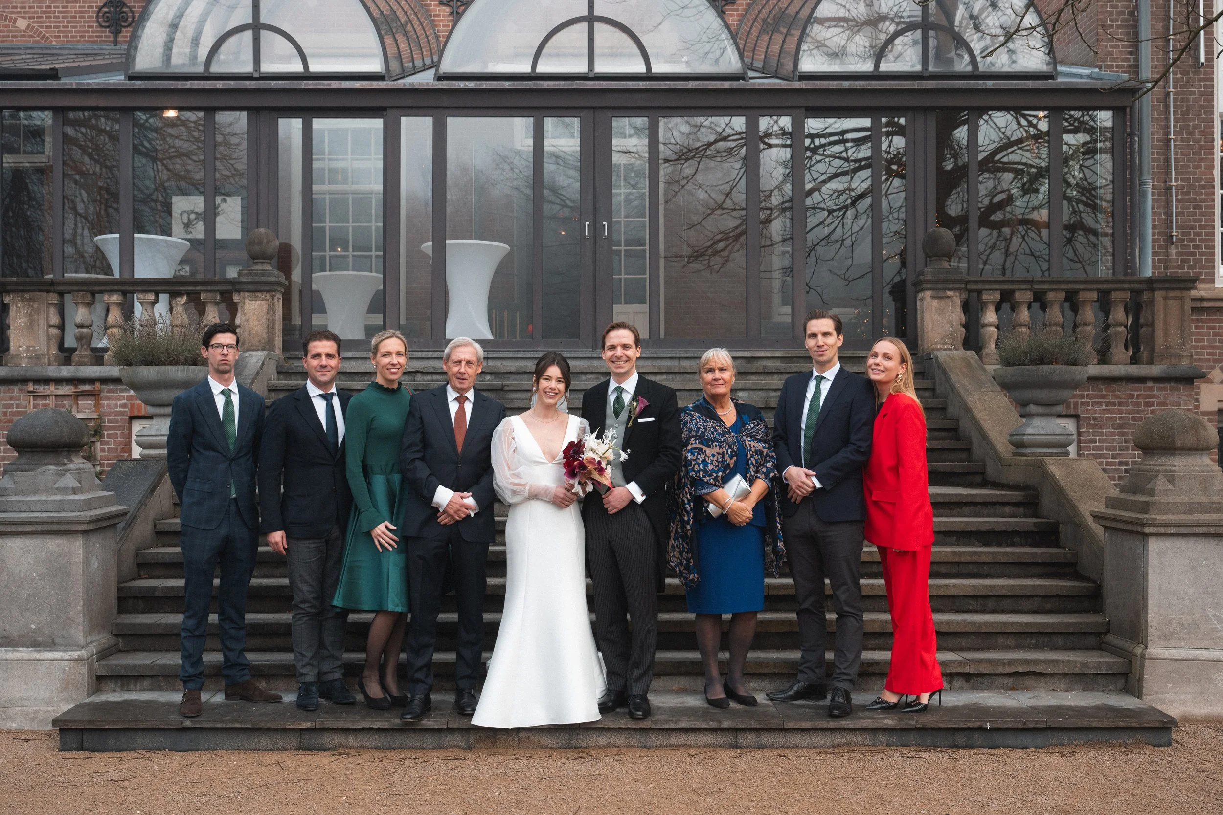 Group of nine people, including a bride and groom, standing on steps in front of a glass conservatory, dressed in formal attire for a wedding.