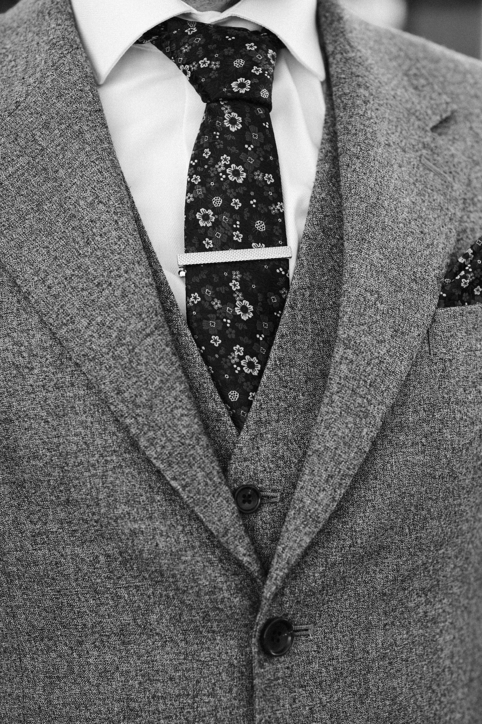 Close-up of a man wearing a gray blazer, a white dress shirt, a floral-patterned tie with a tie clip, and a matching pocket square.