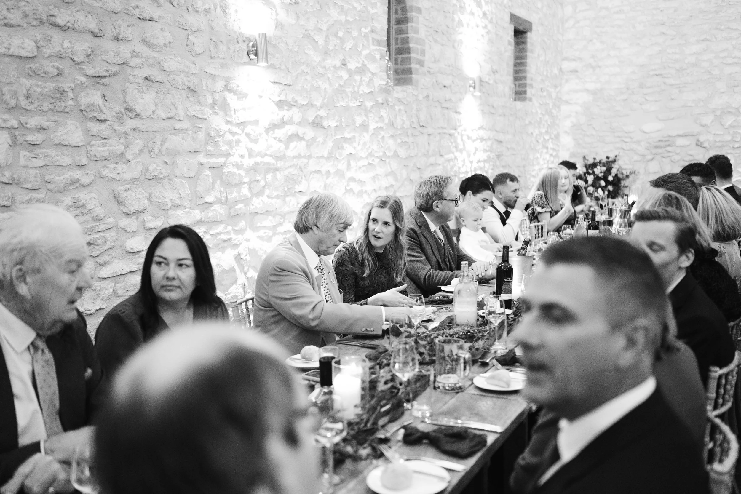 People sitting at a long dining table during a social event or celebration, with a stone wall background and candlelit setting.